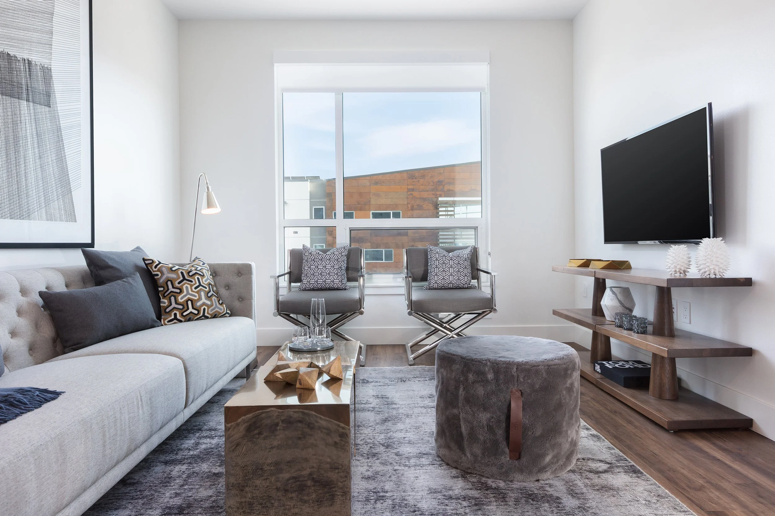 Modern living room with a beige sofa, decorative pillows, a glass top coffee table, two gray chairs with patterned pillows by the window, a flat-screen TV on a wooden console, and a grey ottoman, with a large window and abstract art on the wall.