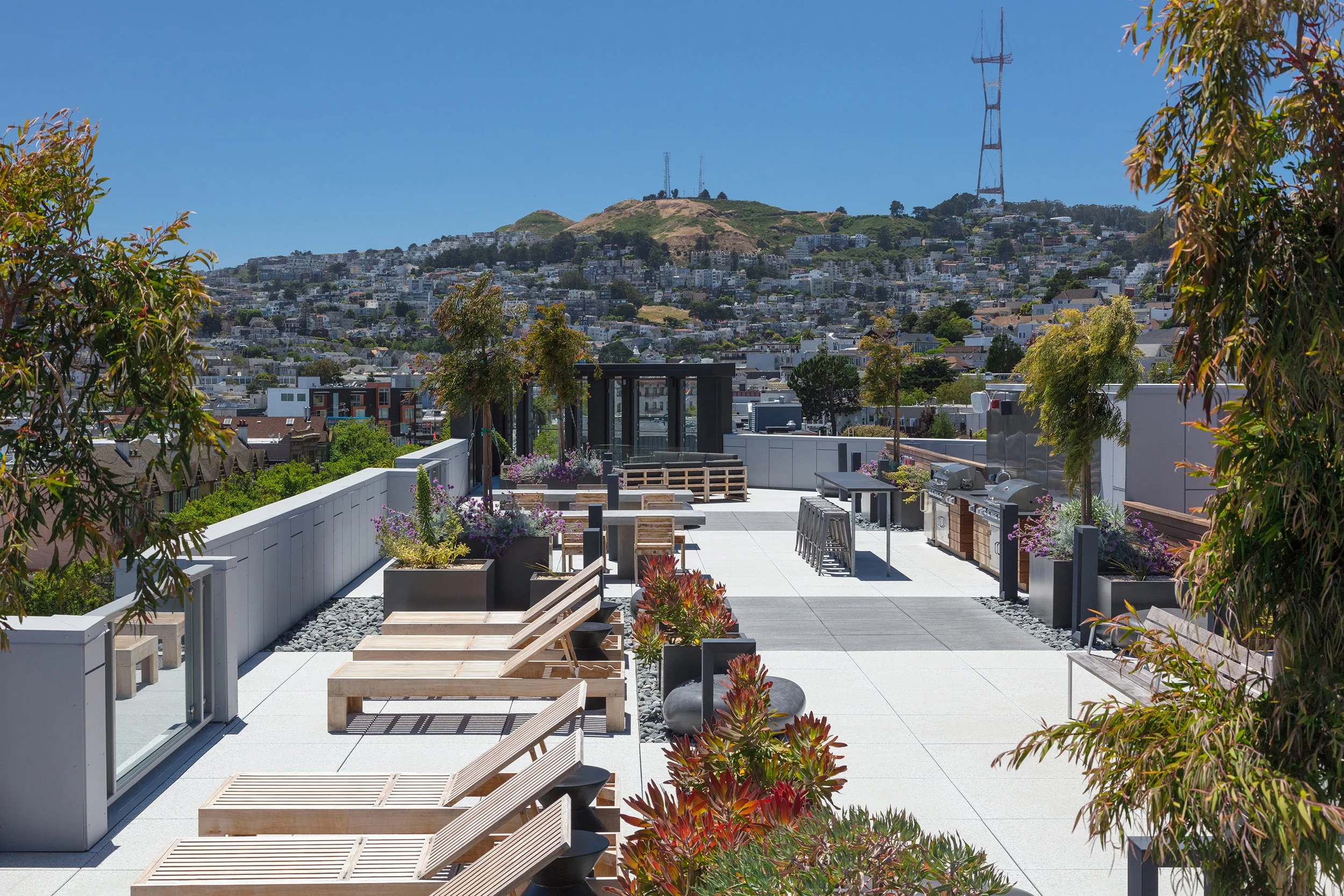 Rooftop terrace with lounge chairs, potted plants, outdoor kitchen, and city view with hills and communication towers in the background on a sunny day.
