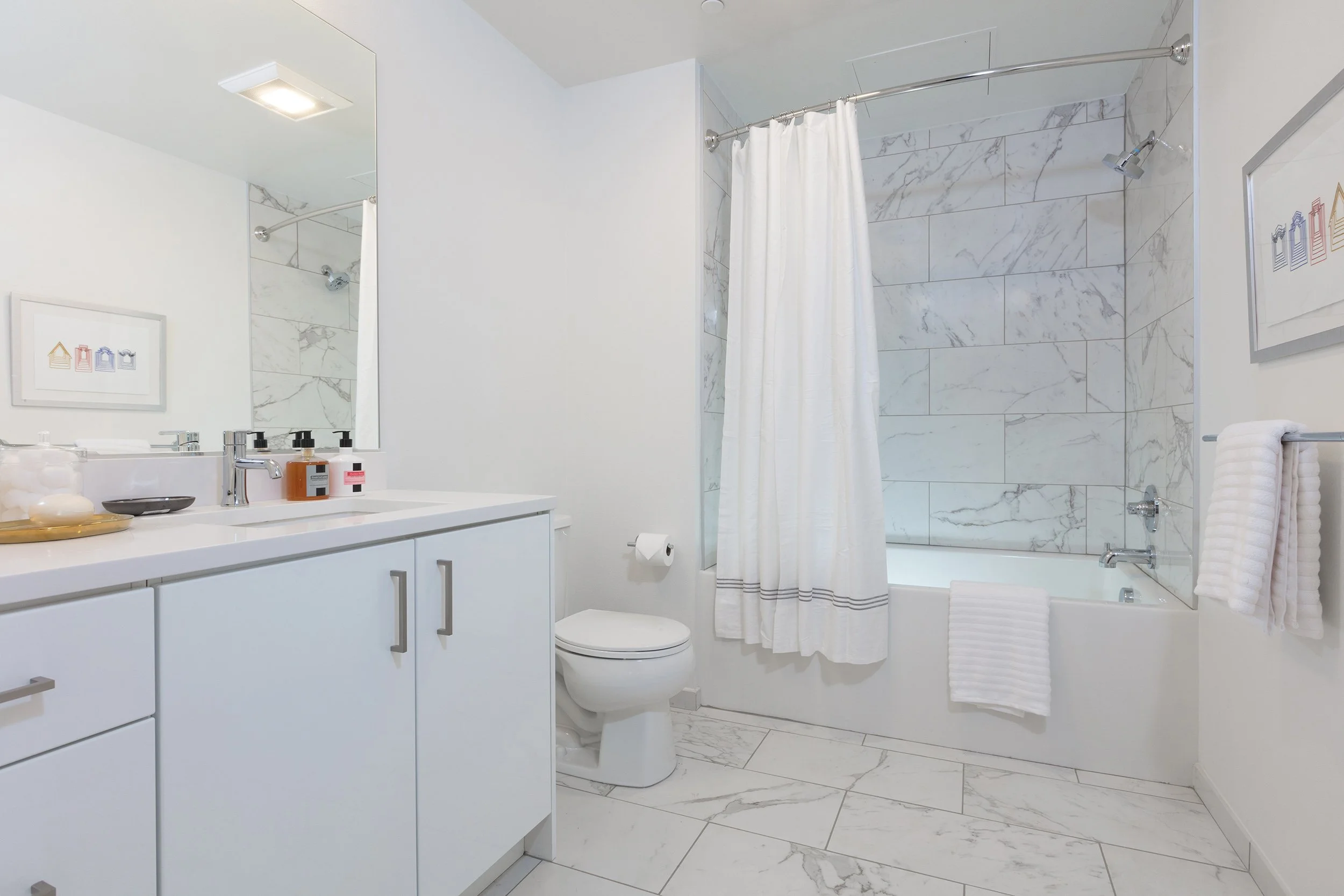 A clean, modern bathroom featuring a white vanity with a mirror, a toilet, a bathtub with a shower, and white towels. The bathroom has marble tiles and minimal decor.