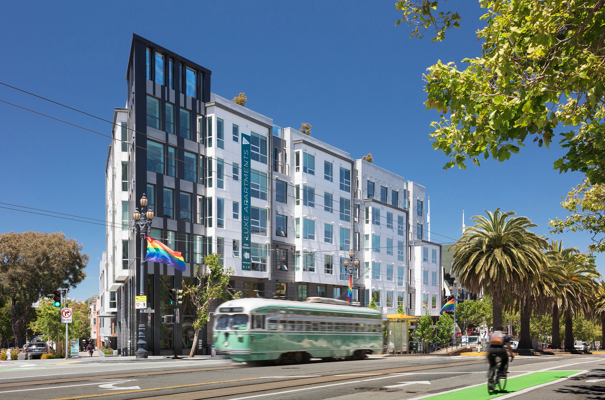 A modern multi-story apartment building with a sign that reads "Luxe Apartments" on a bright, sunny day. There is a green vintage tram and a cyclist on the street, with palm trees and a traffic light in the scene. Pride flags are displayed on lamp posts.