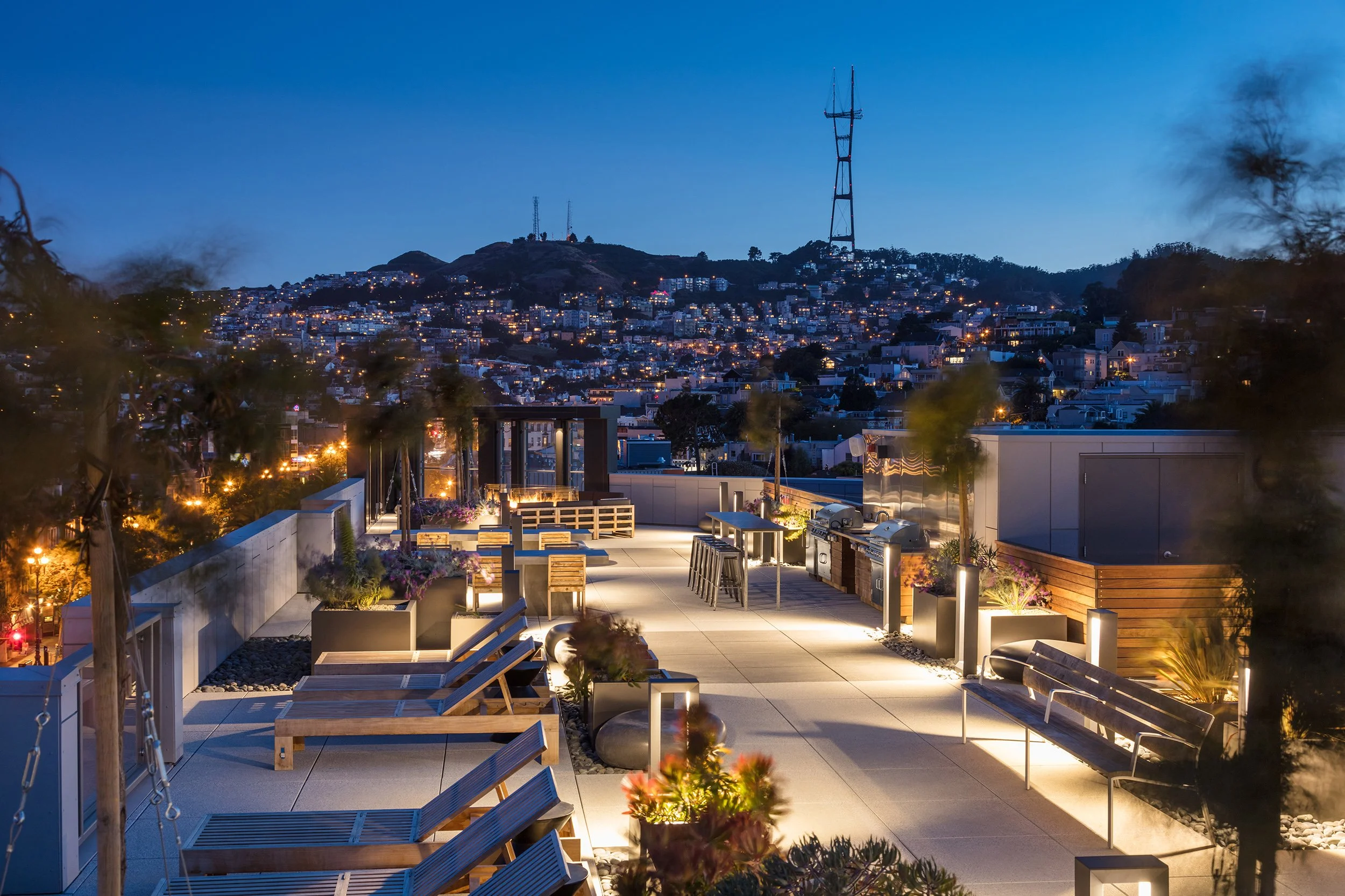 Rooftop patio with lounge chairs, tables, and outdoor kitchen in San Francisco at dusk, city skyline with hilly terrain and communication towers in background