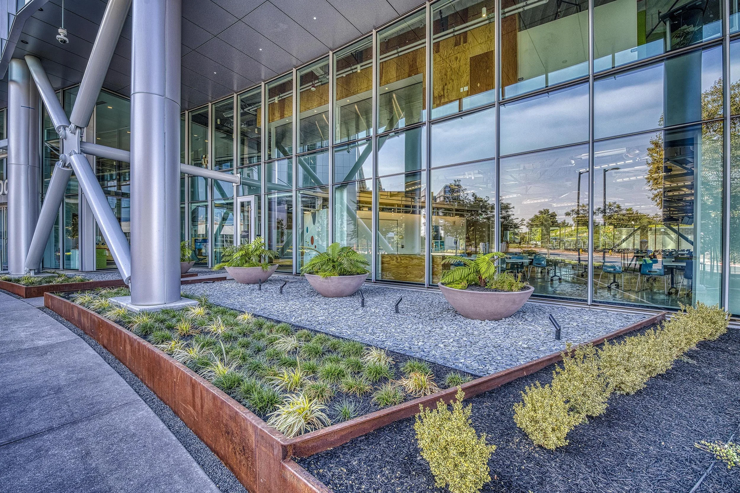 Modern building with glass walls, metal supports, and landscaped outdoor area with plants in planters and flower beds.