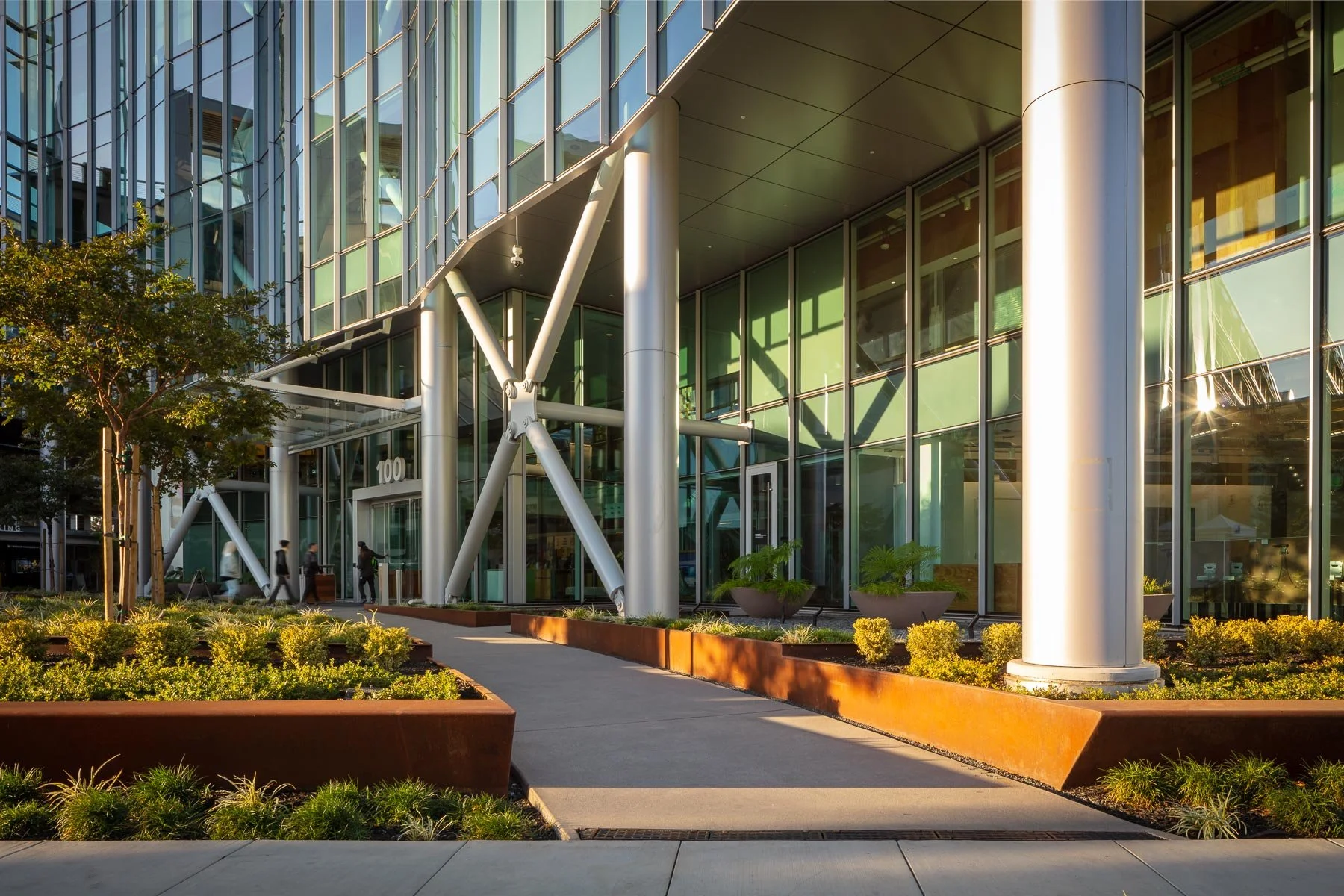 Modern glass office building with large cylindrical white support columns, landscaping with green shrubs and trees, and a sidewalk with pedestrians.