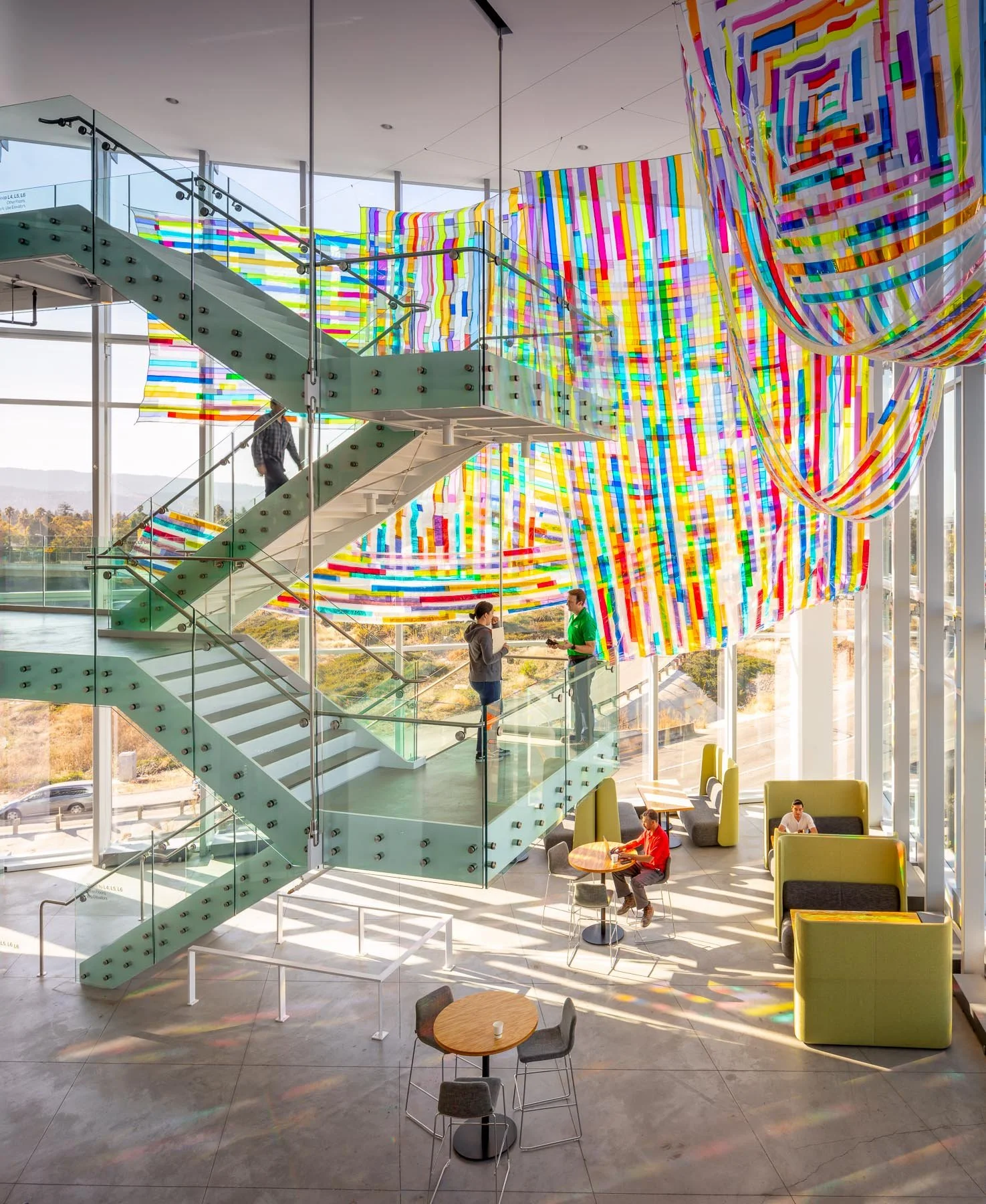 Interior of a modern building with large glass windows, colorful hanging fabric art, a staircase with glass railing, and people sitting and talking.