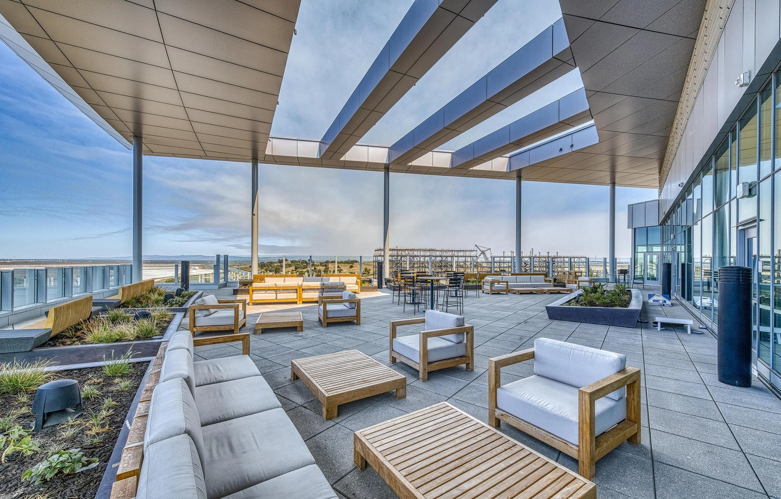Outdoor rooftop terrace with modern furniture, including sofas, armchairs, and tables, under a large canopy with a cityscape and distant mountains in the background.