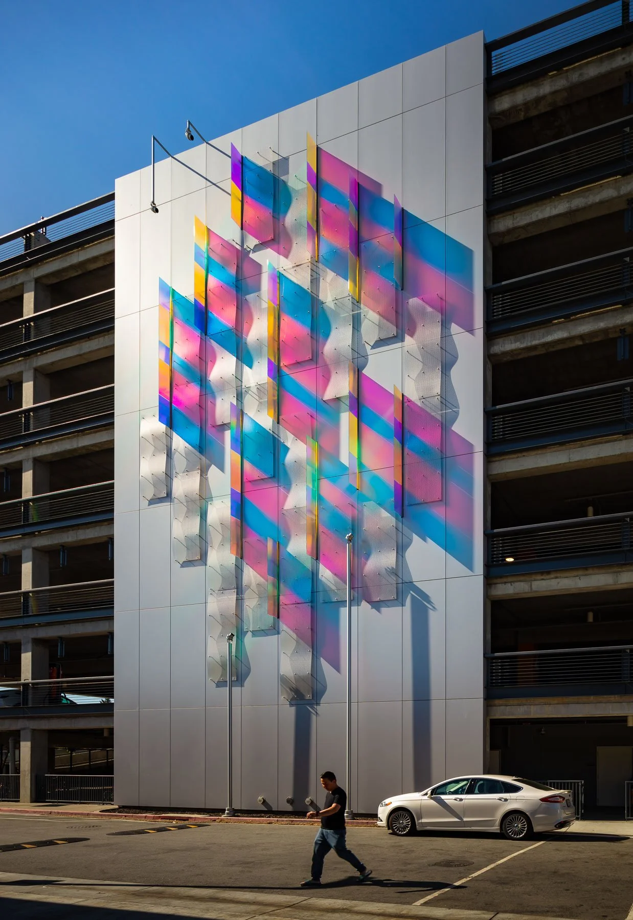 A modern building with a colorful abstract art installation on the exterior wall, featuring pink, blue, and yellow geometric shapes. A man walks in front of a parked white car underneath the artwork.