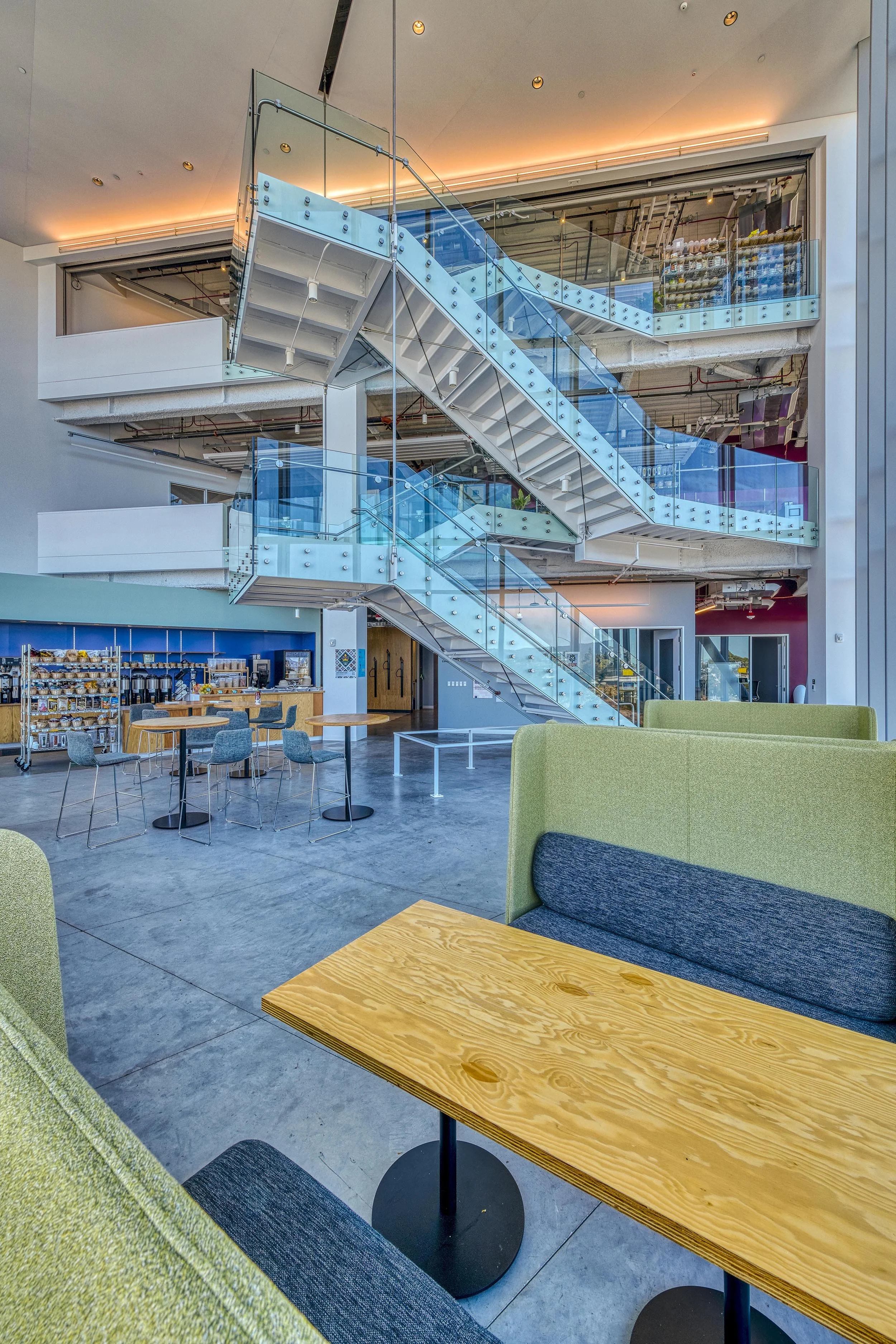 Modern interior of a building with a glass staircase, seating area with green and gray booths, and a coffee station in the background.
