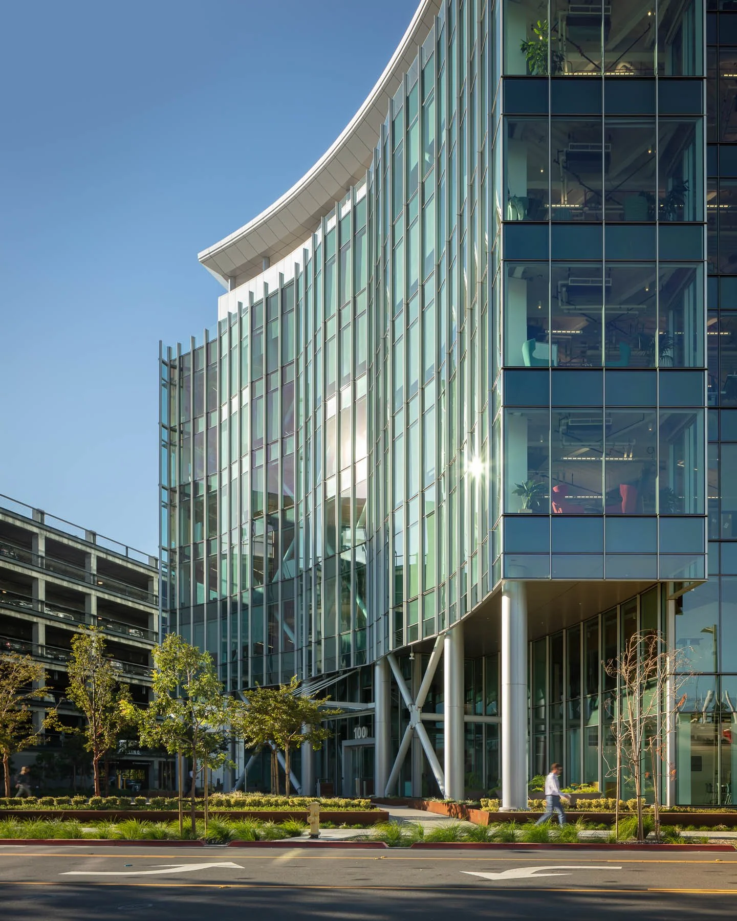 Modern glass office building with trees and a sidewalk in front, sunlight reflecting off the windows.