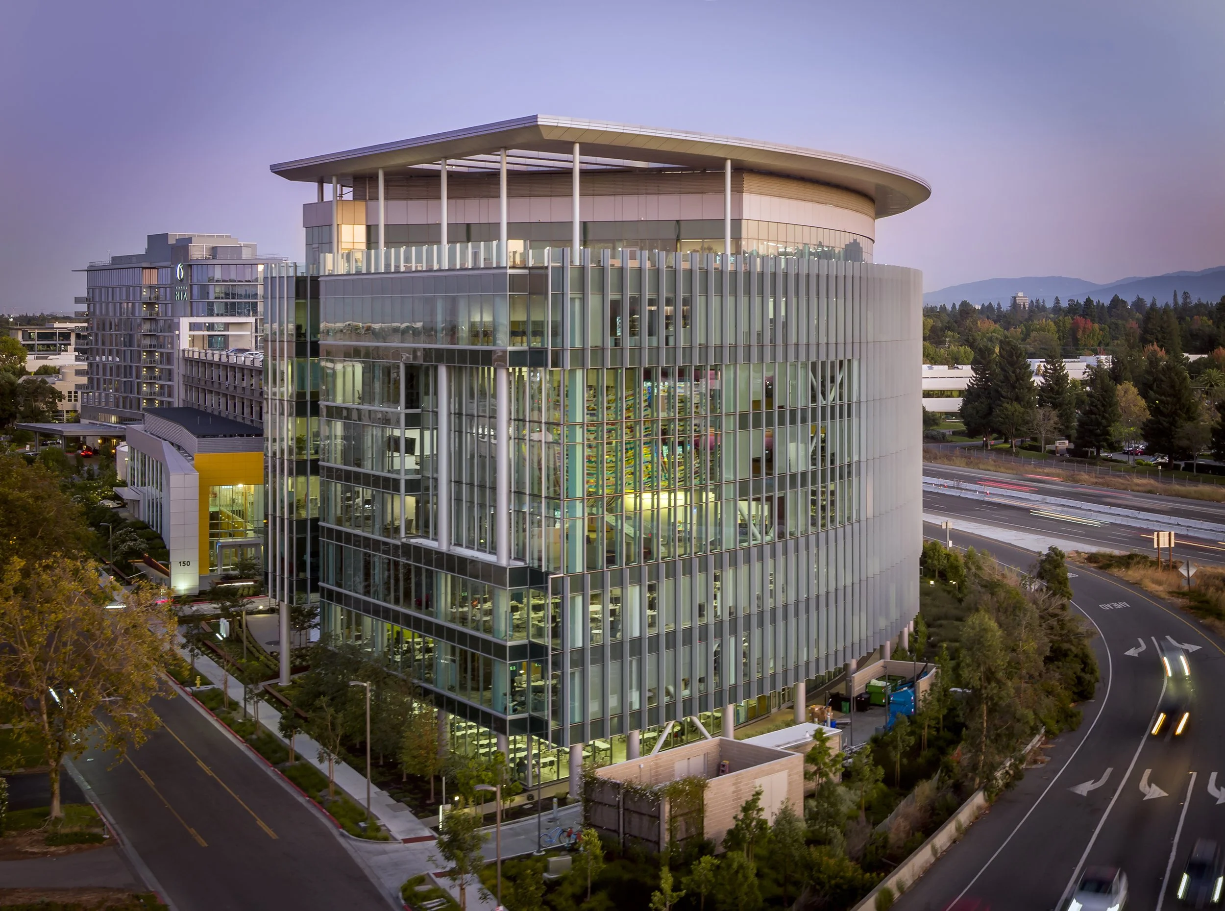 Modern multi-story office building with glass facade, situated near a highway with light traffic, surrounded by trees and greenery, with mountains in the distance at dusk.