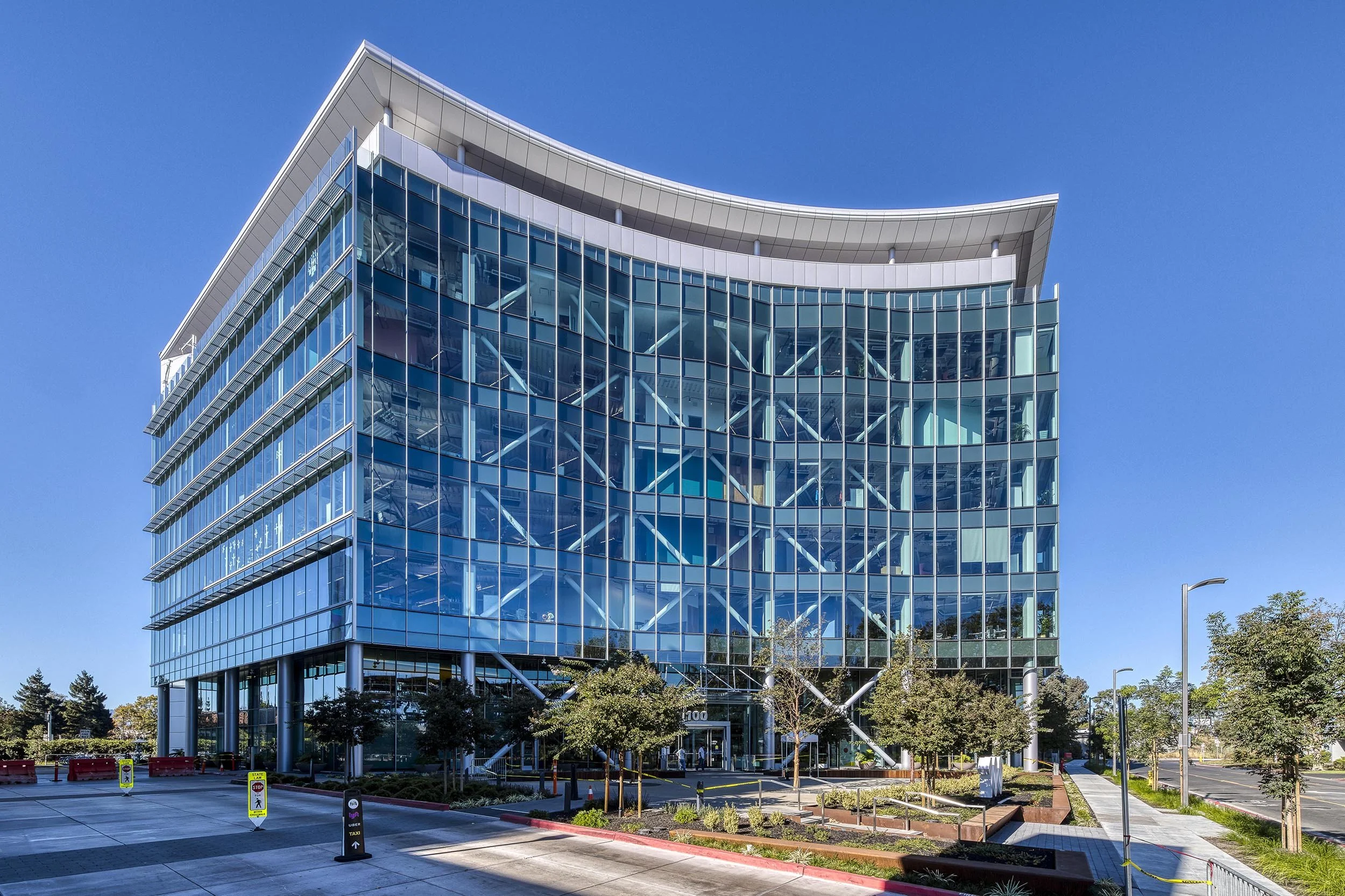 A modern glass office building with a curved roof, several stories tall, with trees and a parking lot in front.
