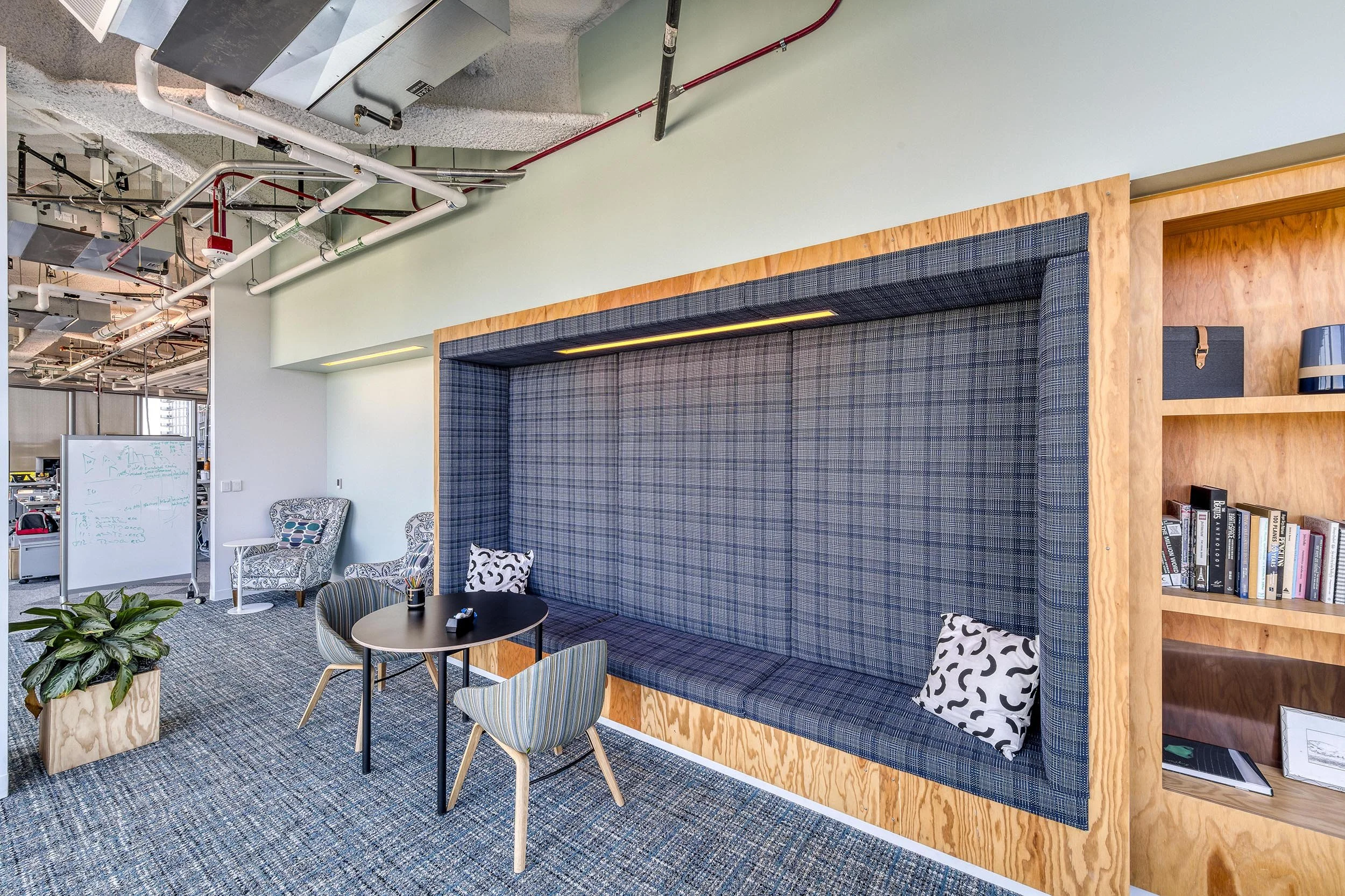 Modern office lounge area with built-in blue plaid fabric seating and decorative pillows, small round table with chairs, potted plant, bookshelves, and an open office space with a whiteboard and overhead wiring.