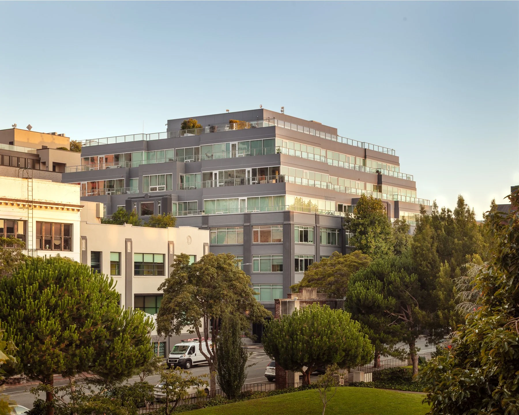 Multi-story residential building with glass balconies, surrounded by trees and parked cars, under a clear sky.