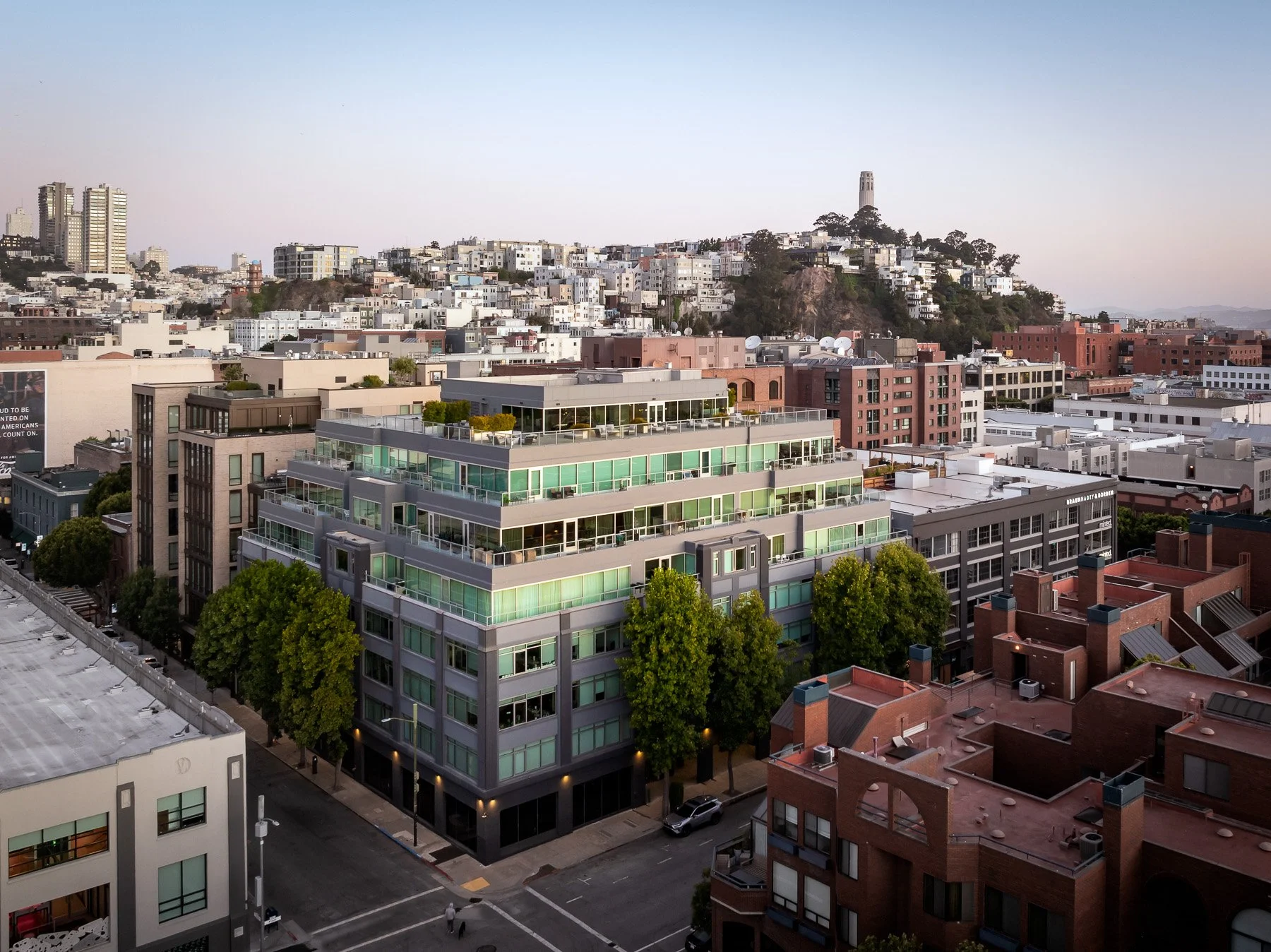 A cityscape with a modern multi-story building in the foreground, surrounded by trees, and various buildings including high-rises and a hill with houses and a landmark tower in the background.