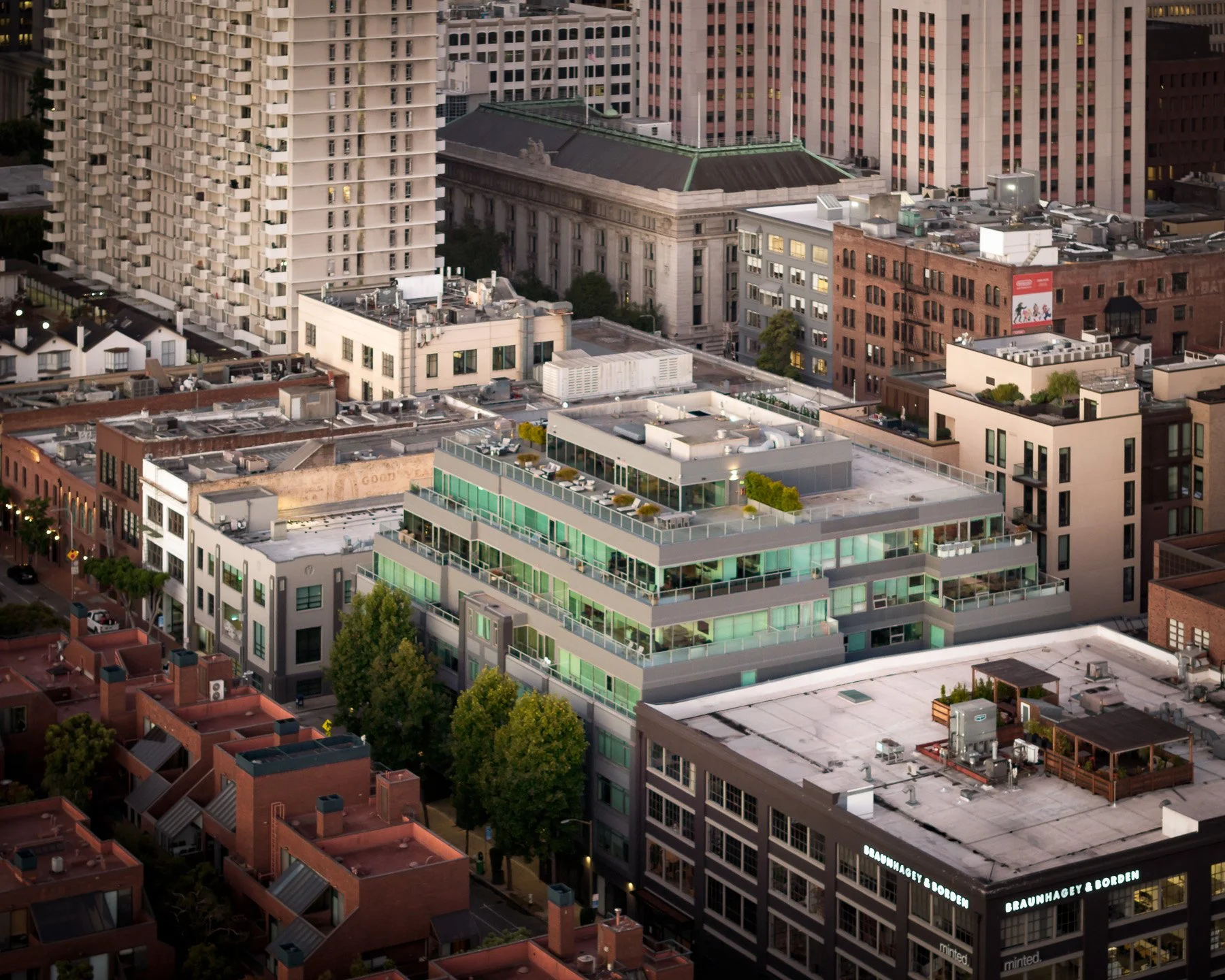 Aerial view of city buildings including a modern multi-story building with green-tinted glass balconies and trees surrounding the area.