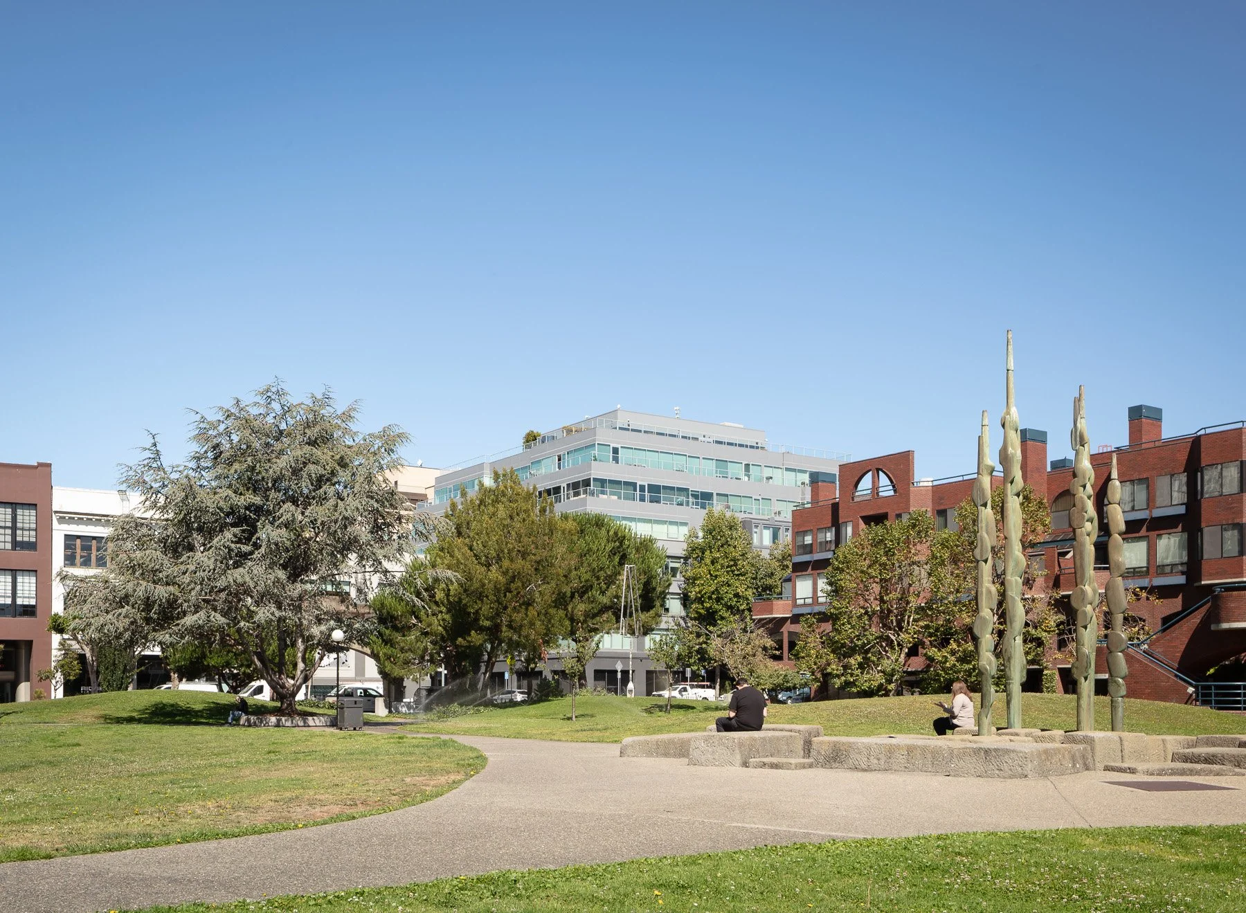 Urban park scene with trees, grass, and sculptures, surrounded by modern buildings, on a sunny day.