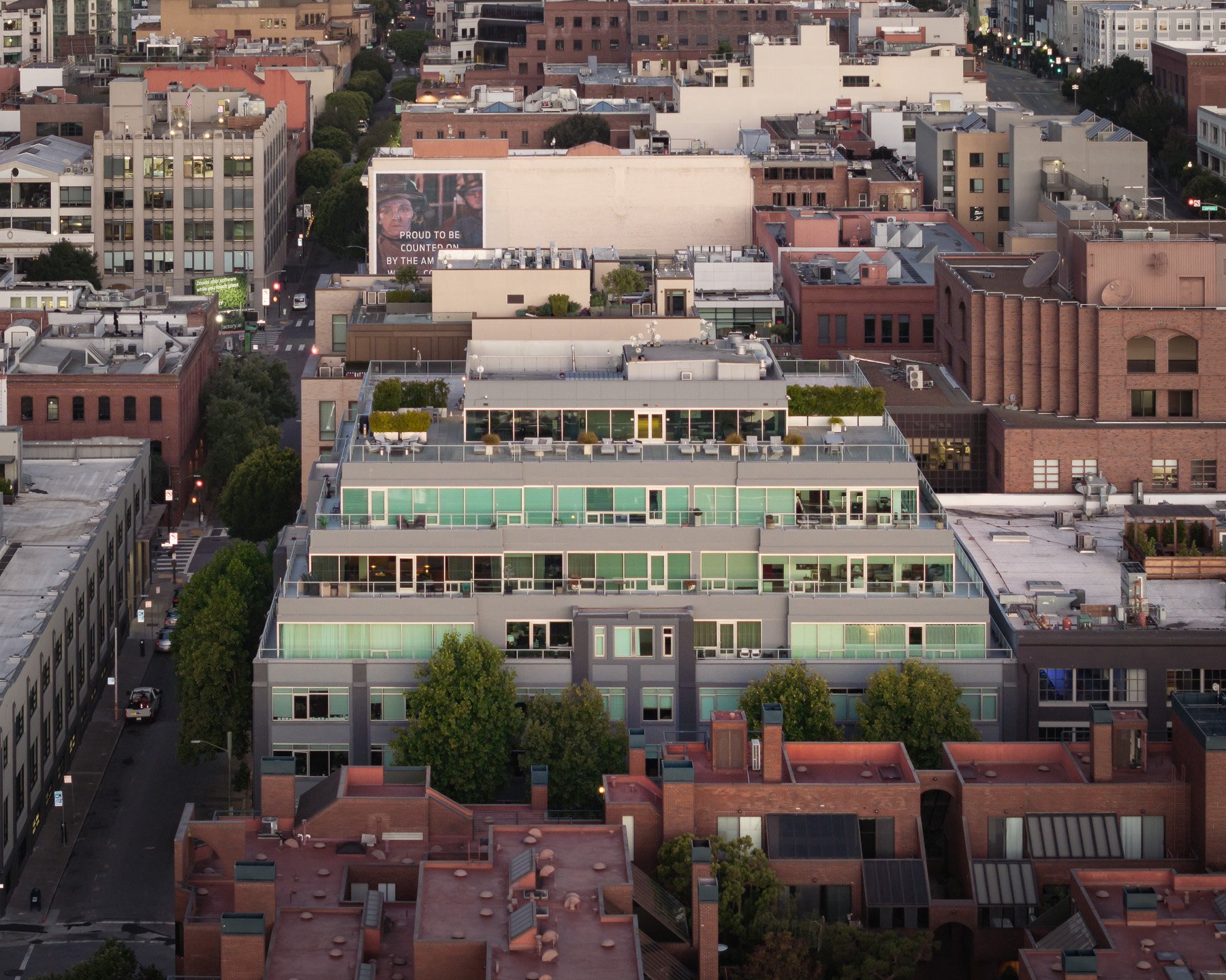 Aerial view of a city block with a modern apartment building featuring multiple balconies and rooftop terraces, surrounded by older brick and concrete buildings, with streets and trees visible.