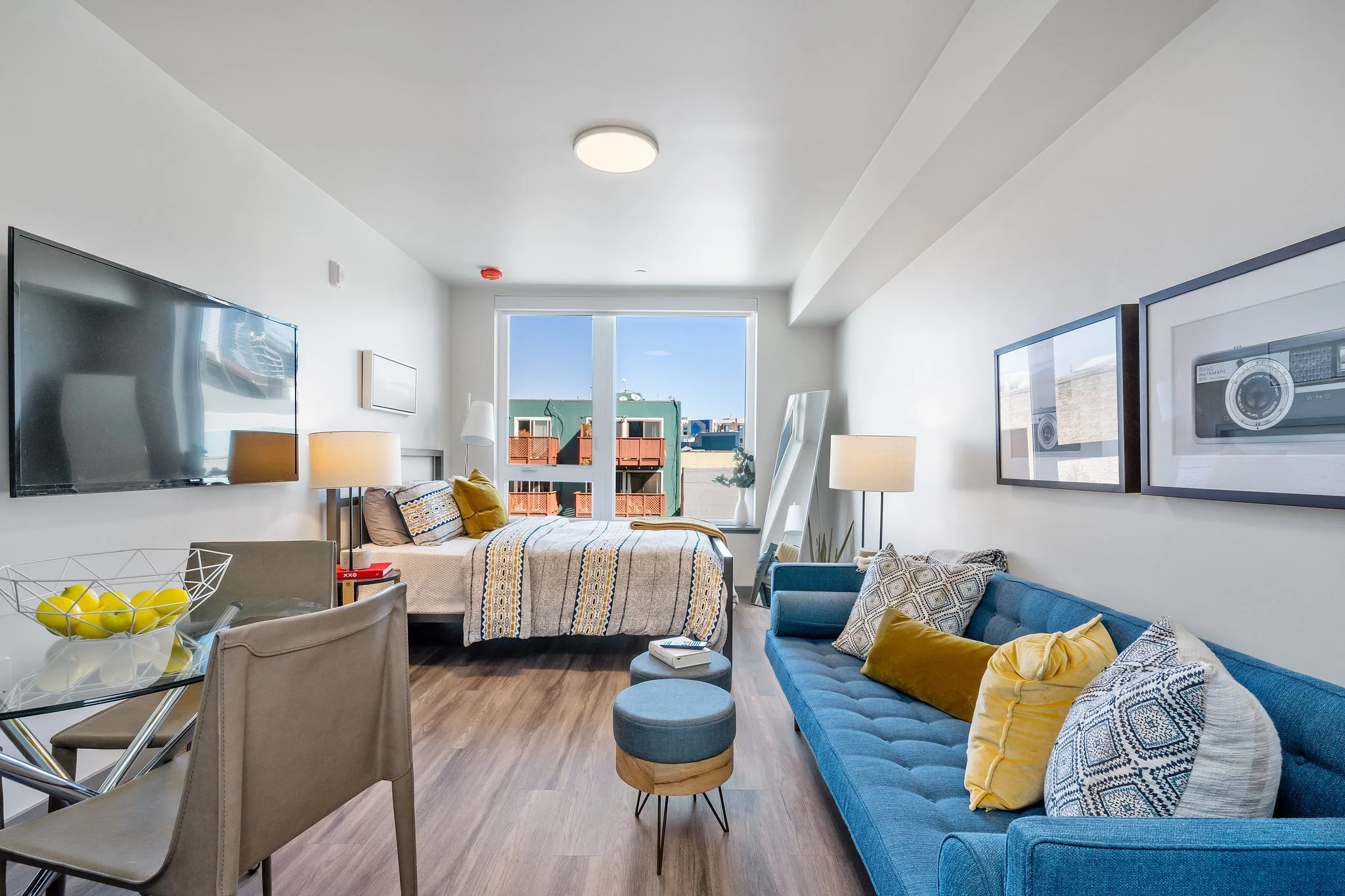 Modern apartment room with a bed near the glass door, a blue sofa with yellow and patterned pillows, a glass dining table with chairs, framed artwork, and a balcony view of neighboring buildings