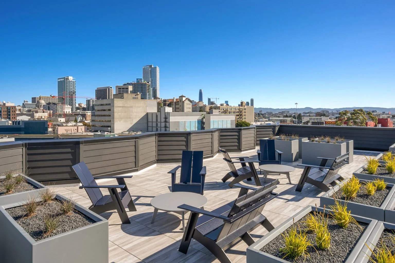 Rooftop terrace with modern outdoor chairs, tables, and planters, overlooking a city skyline with tall buildings under a clear blue sky.