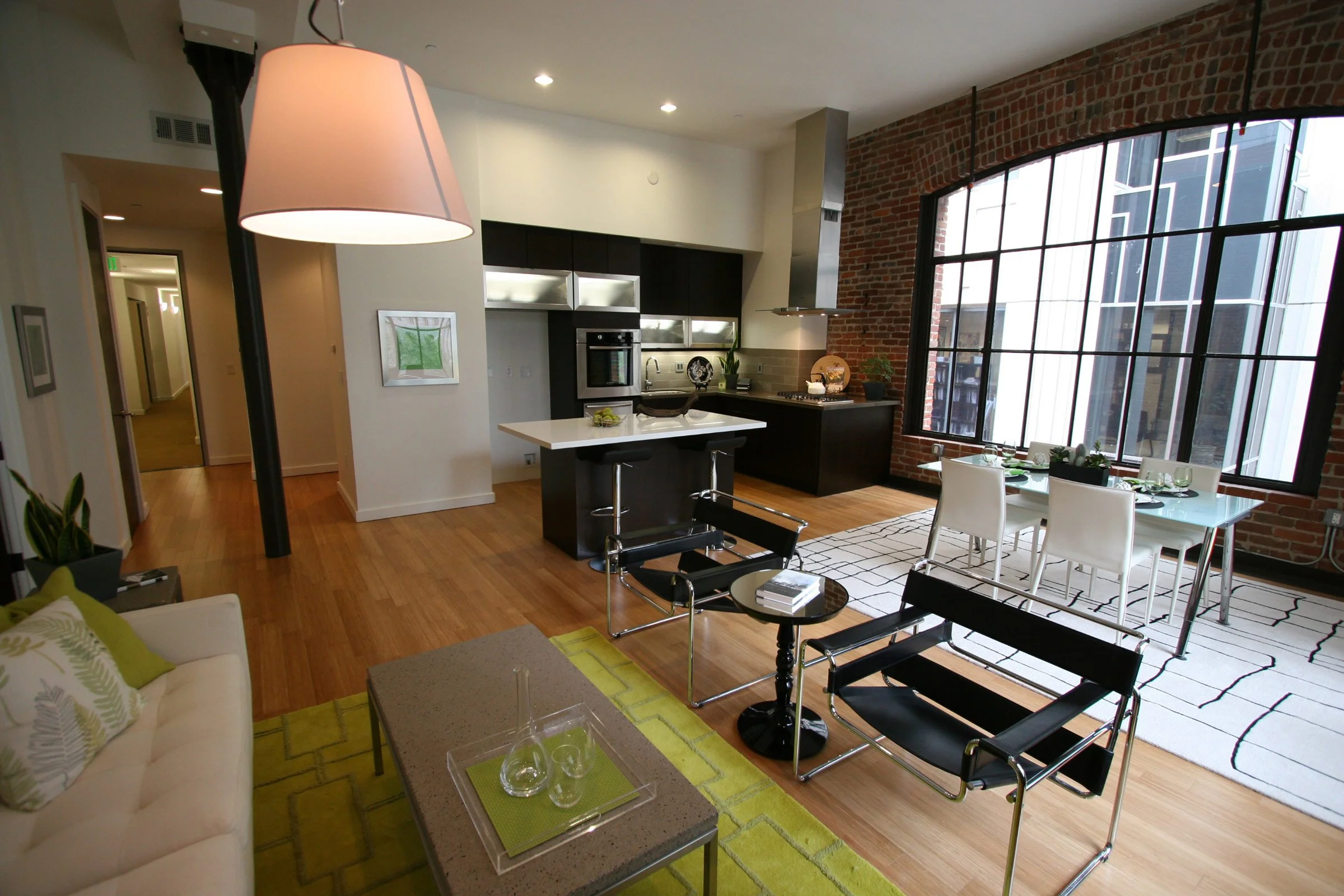 Open-concept living room and kitchen with exposed brick walls, large windows, wooden floors, modern furniture, and a dining table.