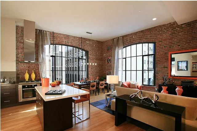 Living room and kitchen area with large windows, brick walls, and modern furniture.
