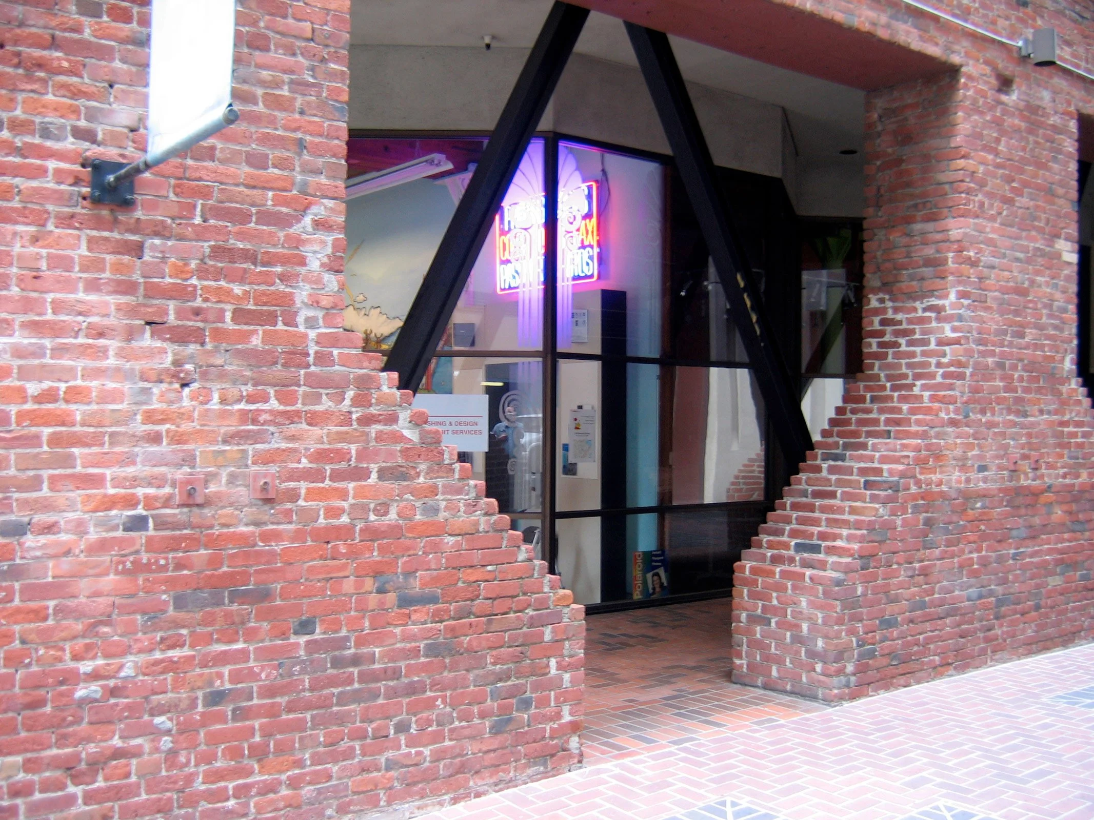 Part of a brick building with a storefront window, showing a neon sign that reads 'Pasta & Pastries' inside, with architectural black support beams around the window opening.
