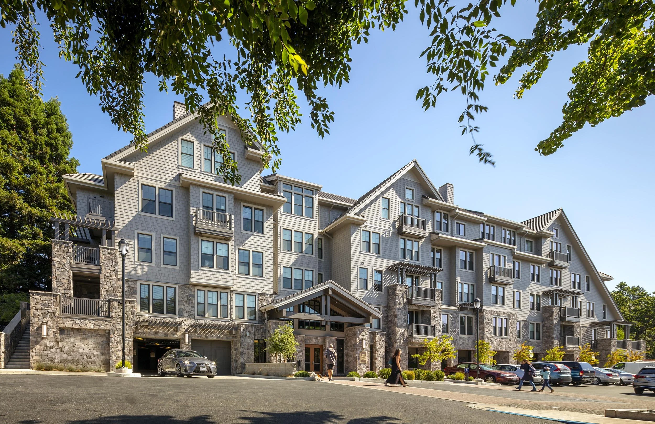 Multi-story residential apartment building with stone and light-colored siding, multiple balconies, and surrounded by a parking lot with cars and pedestrians.