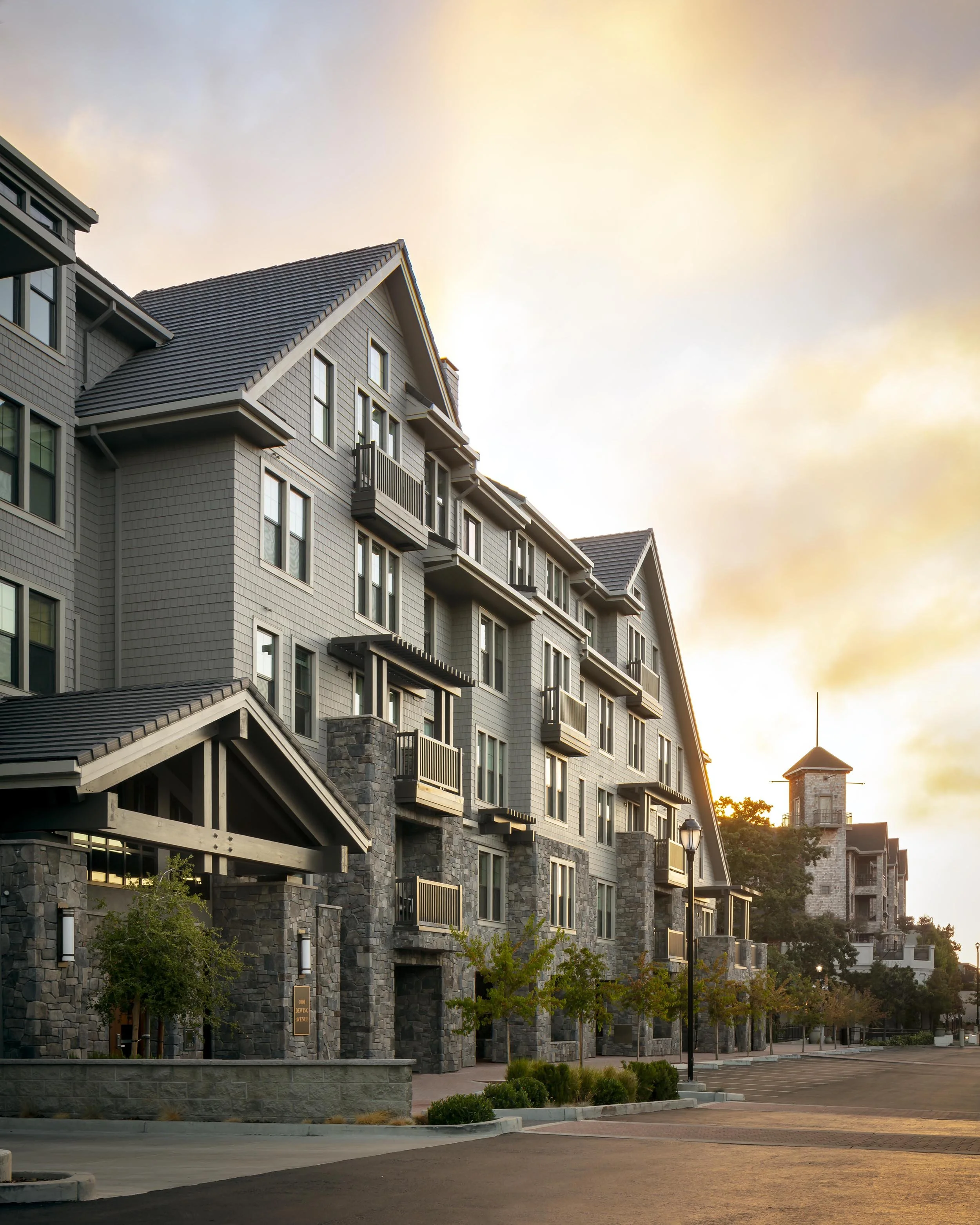 Multi-story residential building with stone accents and small balconies, lit by warm sunset light, with a paved street and trees in front.