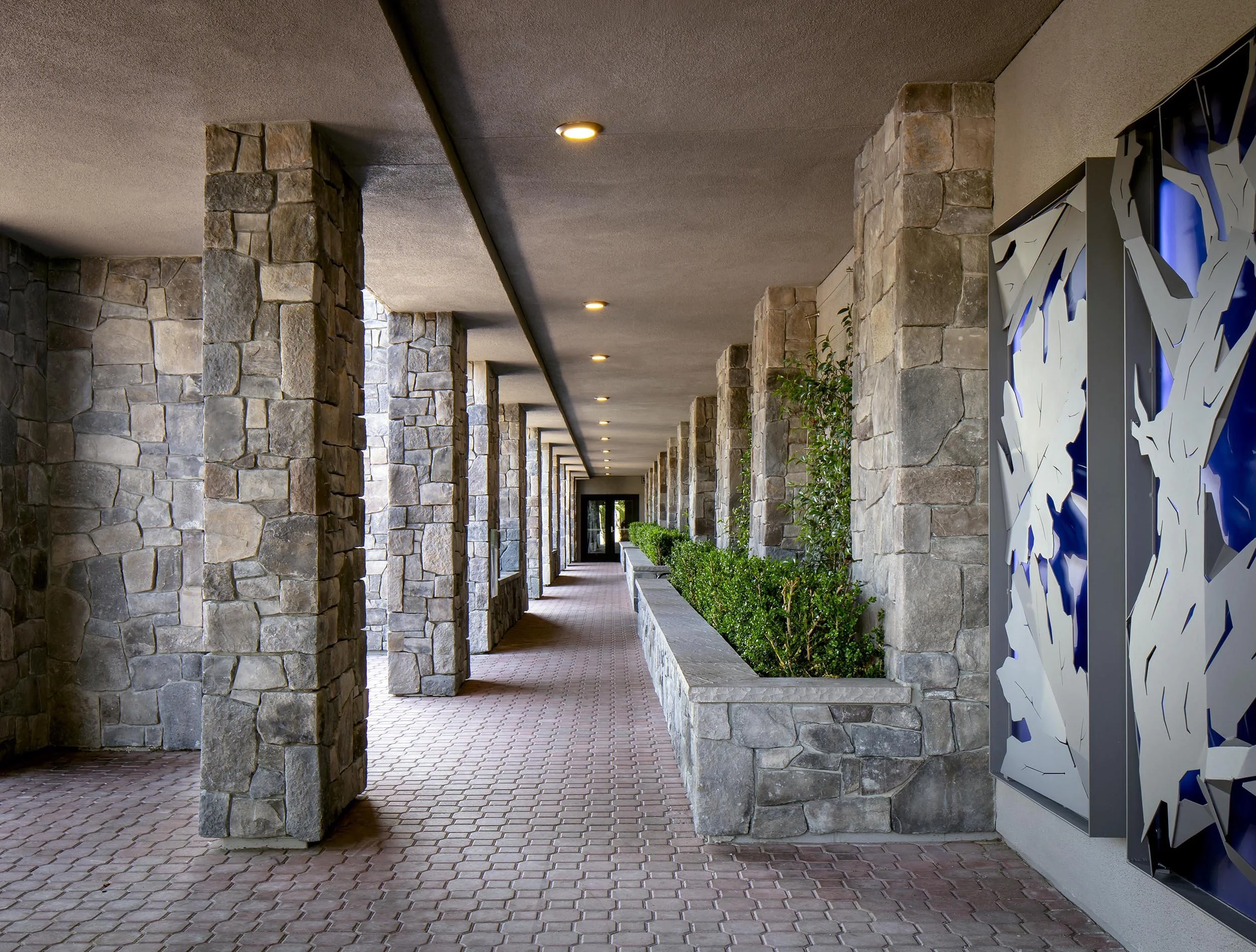 Covered outdoor walkway with stone columns and walls, brick floor, and decorative panel on the right side with abstract design and blue accents.