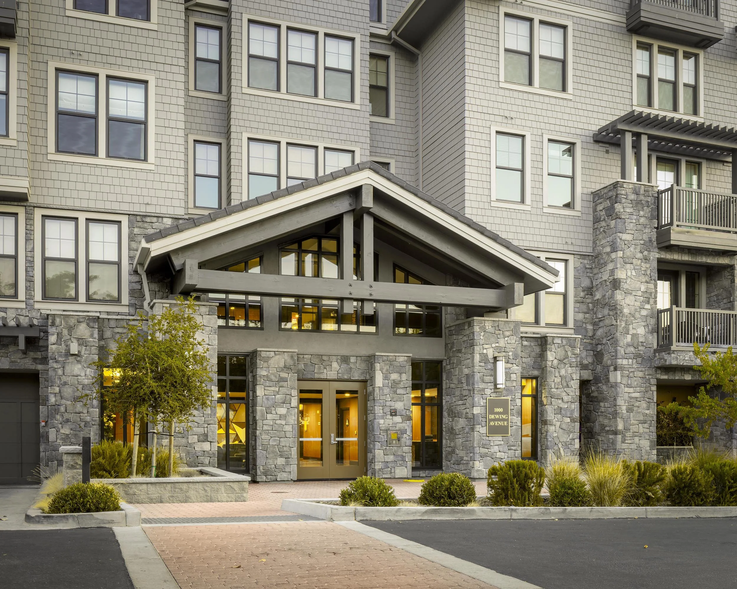 Front entrance of a modern multi-story apartment or condominium building with stone and siding exterior, glass doors, and multiple windows.