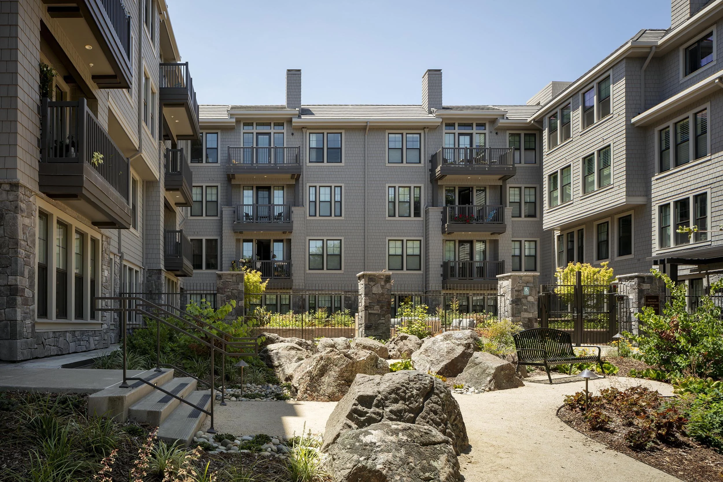 A multi-story apartment building with balconies, gray exterior, and numerous windows, surrounding a landscaped courtyard with rocks, plants, a bench, and a gravel pathway.