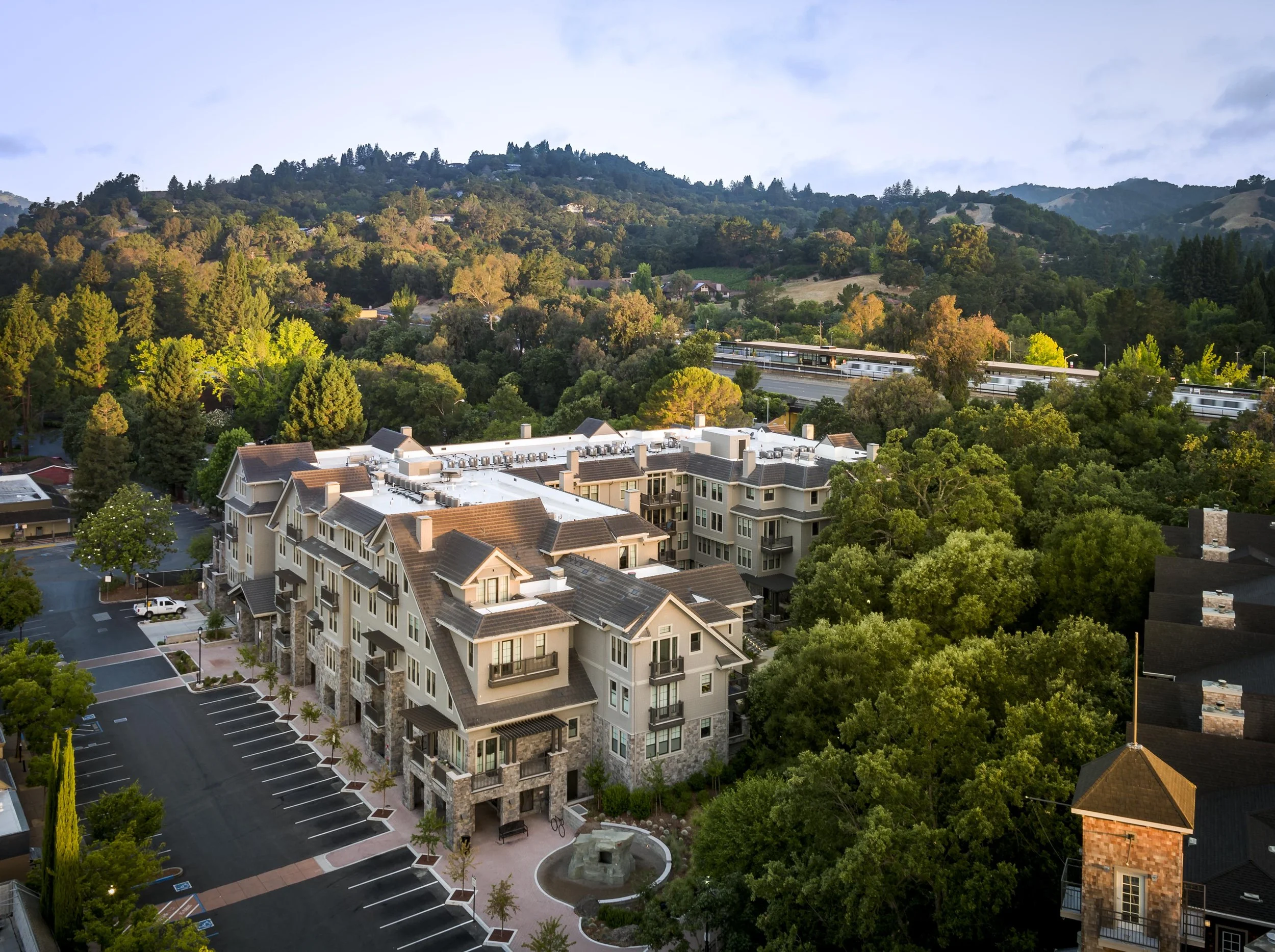 An aerial view of a residential area with apartment buildings surrounded by lush green trees, with hills in the background and a train track in the distance.