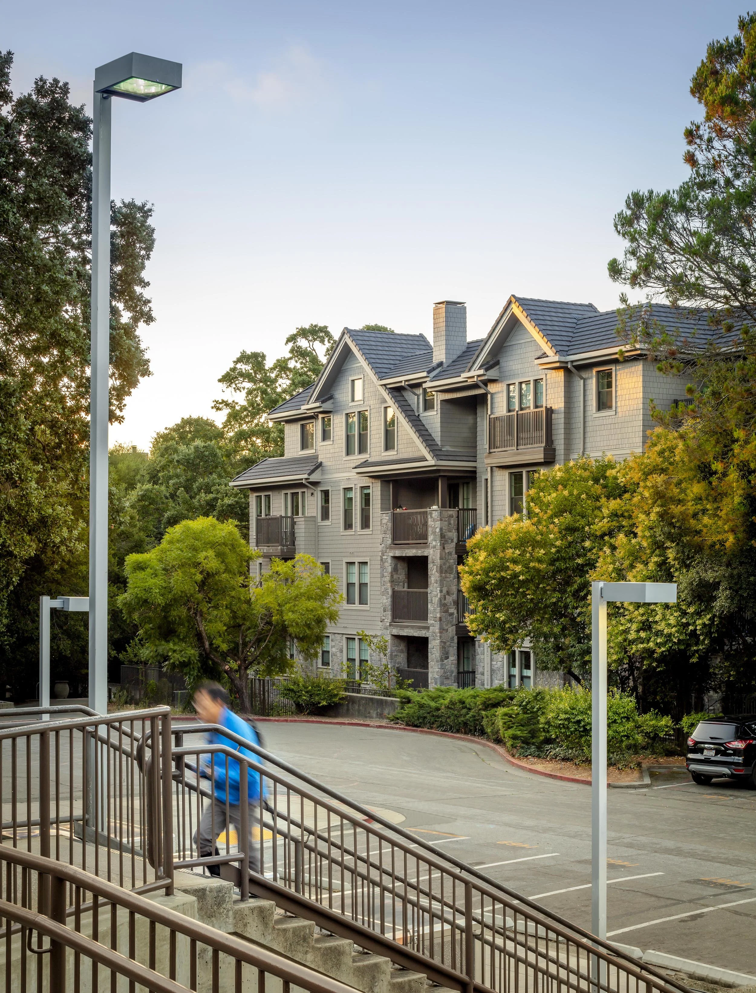 A person in a blue jacket walking down outdoor stairs in parking lot with residential apartment building, trees, and streetlights in background.