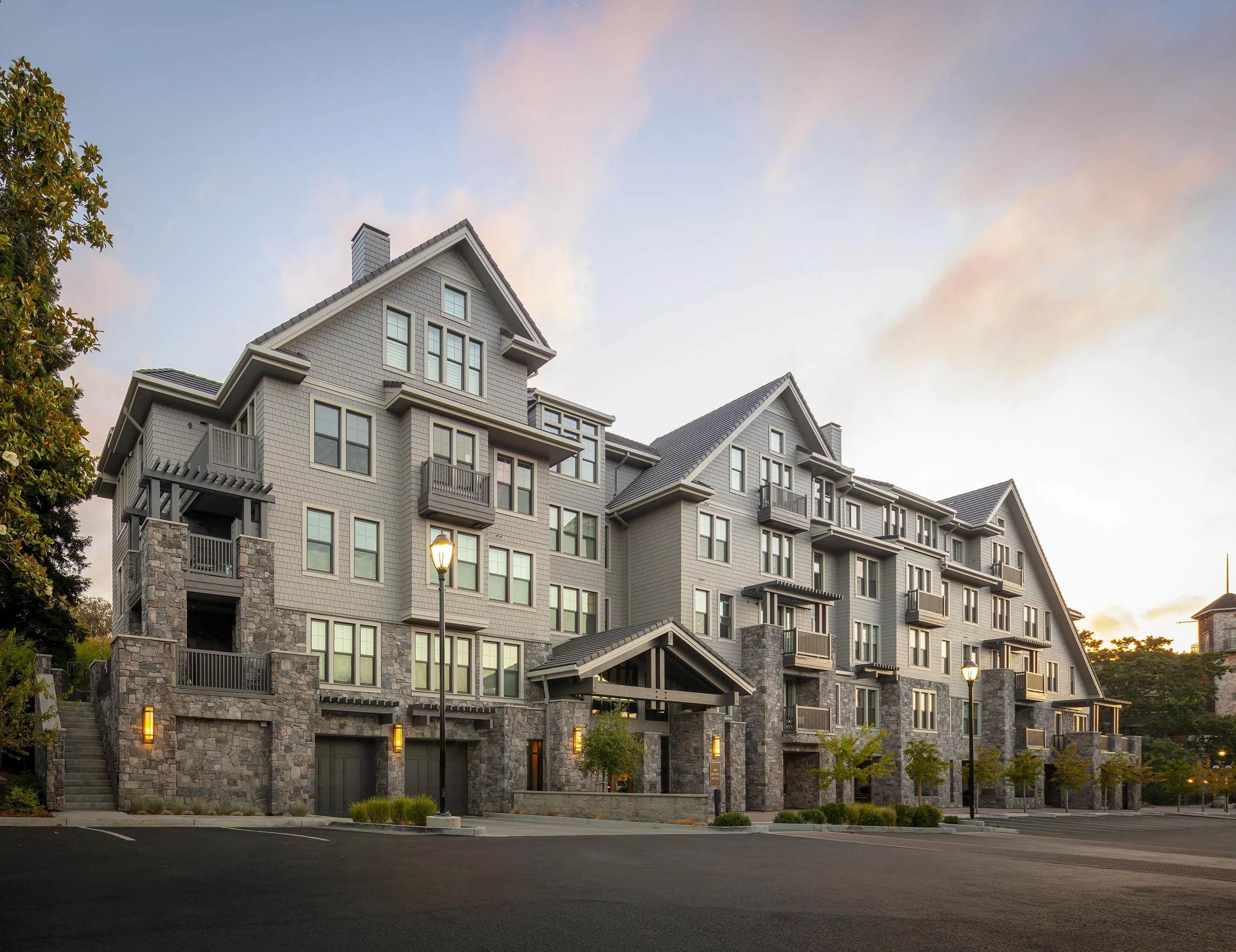 Upscale apartment building with stone accents, multiple balconies, and large windows, taken at sunset with a clear sky and street lamps in front.