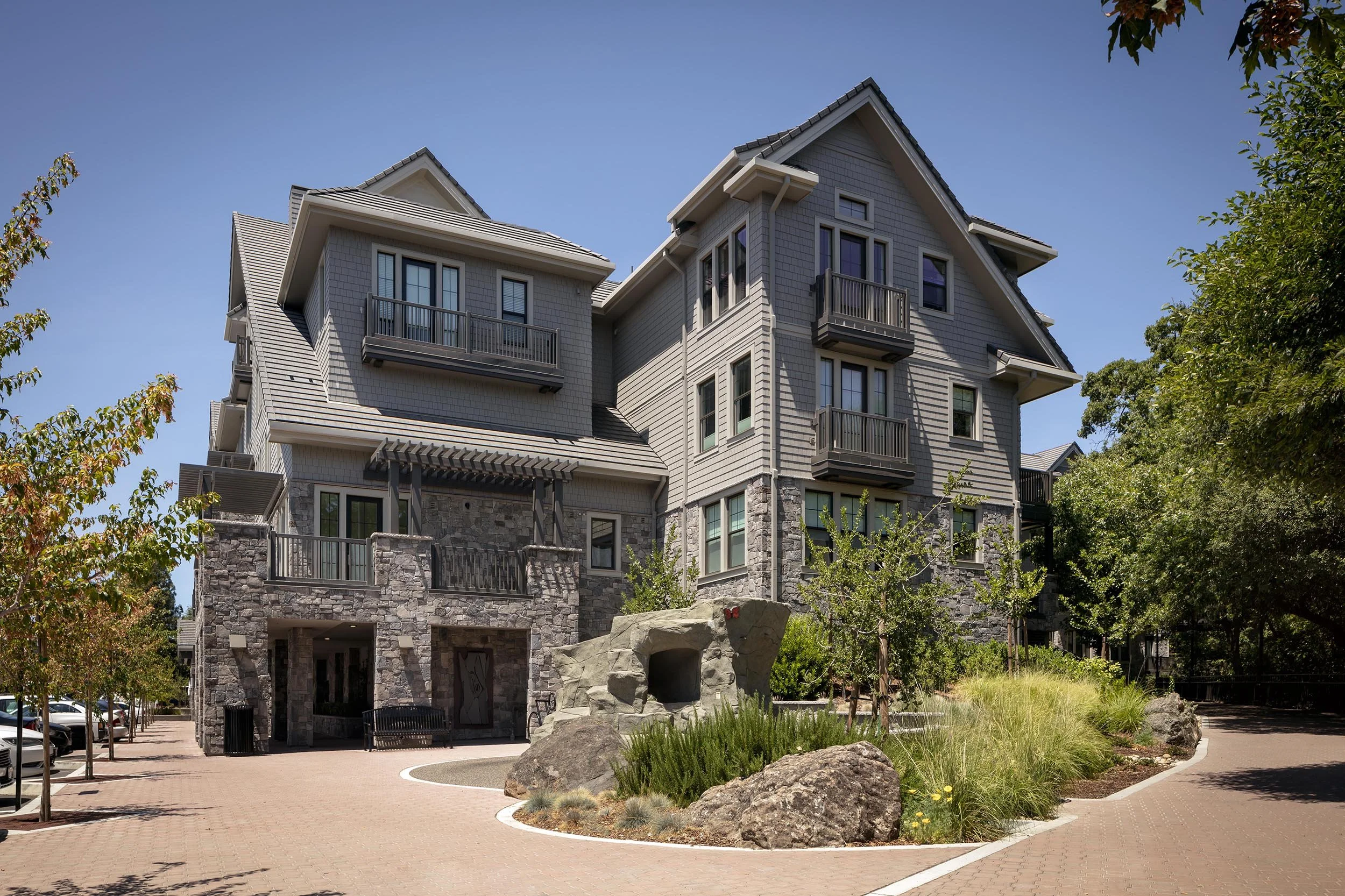 Multi-story residential building with gray siding, stone accents, balconies, and a landscaped front area with rocks, trees, and plants.