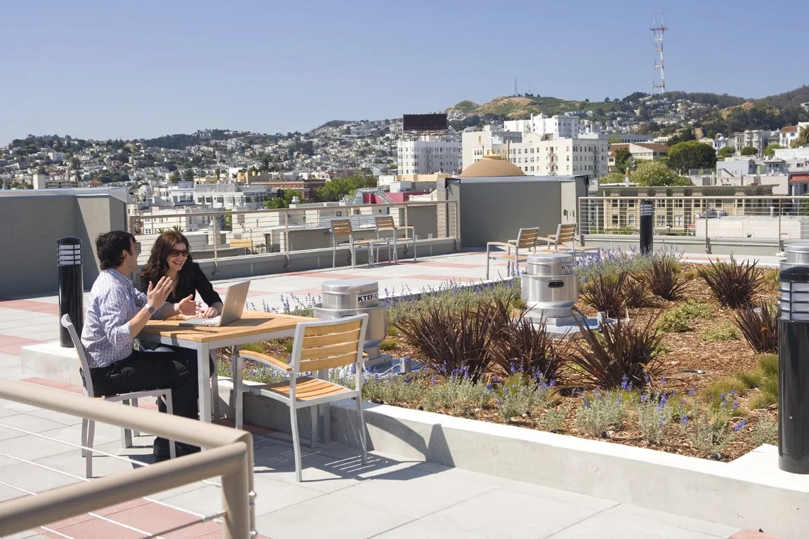 Two people sitting at outdoor table on a rooftop, working on a laptop and talking, with cityscape and hills in the background.