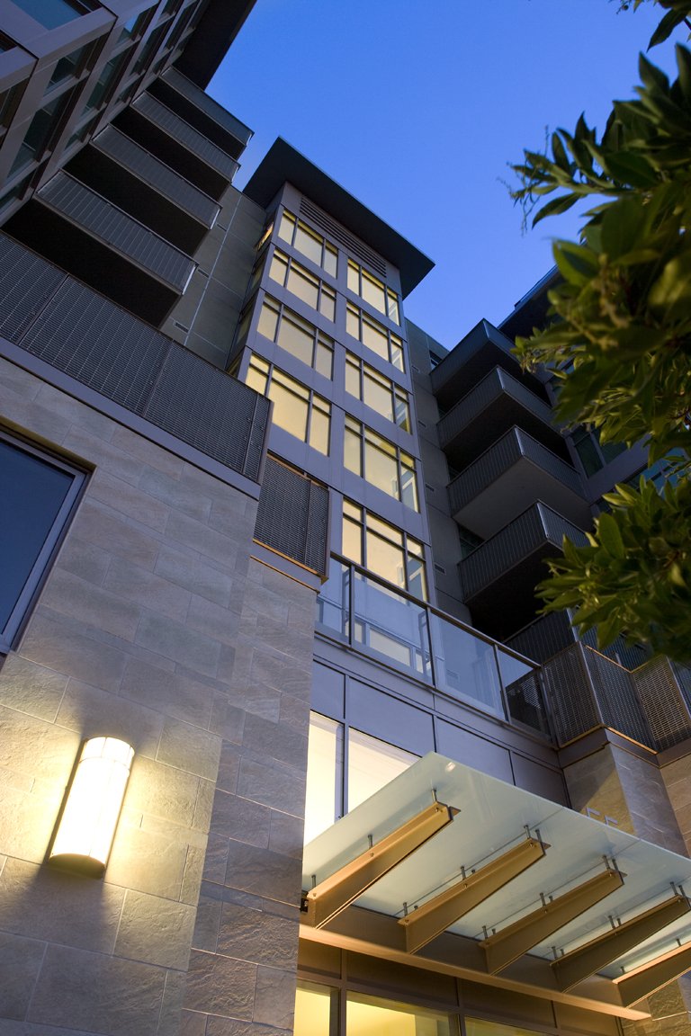 Low-angle view of a modern apartment building with glass balconies and large windows at dusk, with a tree partially visible on the right and a wall-mounted light on the left.