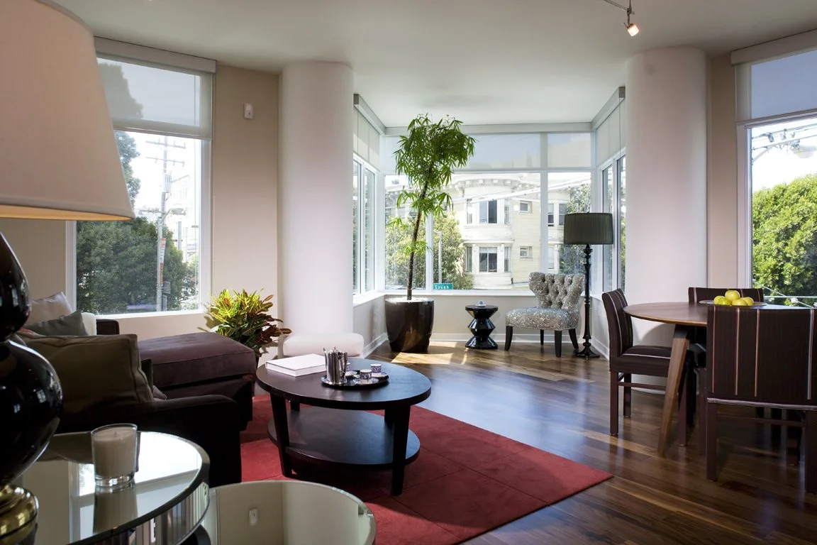 Bright living room with large windows, hardwood floors, a dark sofa, a round coffee table, potted plant, upholstered armchair, floor lamp, and dining table with apples.