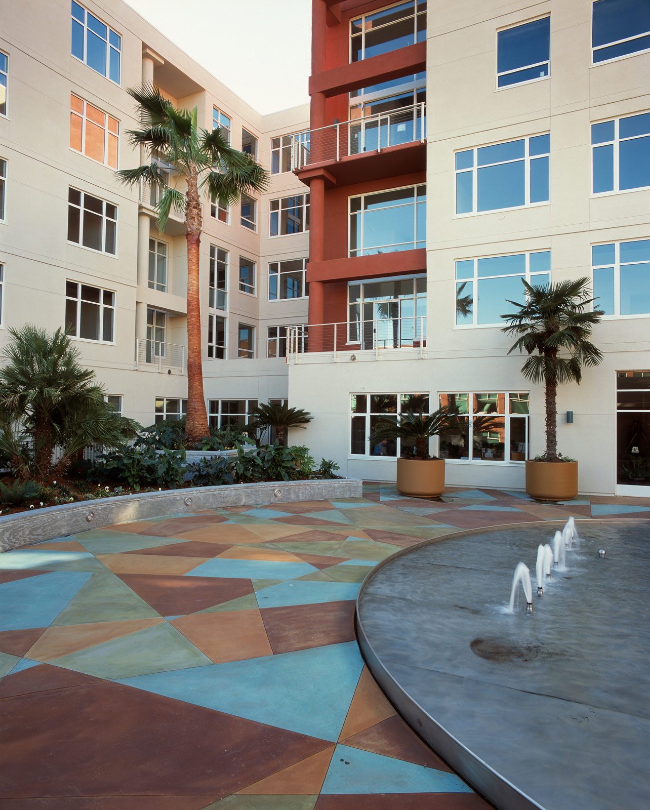 Interior courtyard of a modern apartment complex with colorful geometric patterned flooring, palm trees in large pots, and a small fountain with water jets.