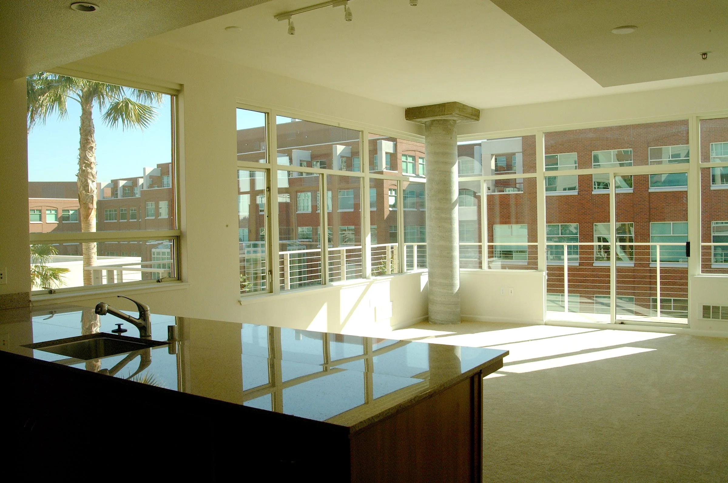 Empty apartment with large floor-to-ceiling windows and a kitchen counter with a sink, sunlight streaming in, and a view of other modern apartment buildings and palm trees outside.