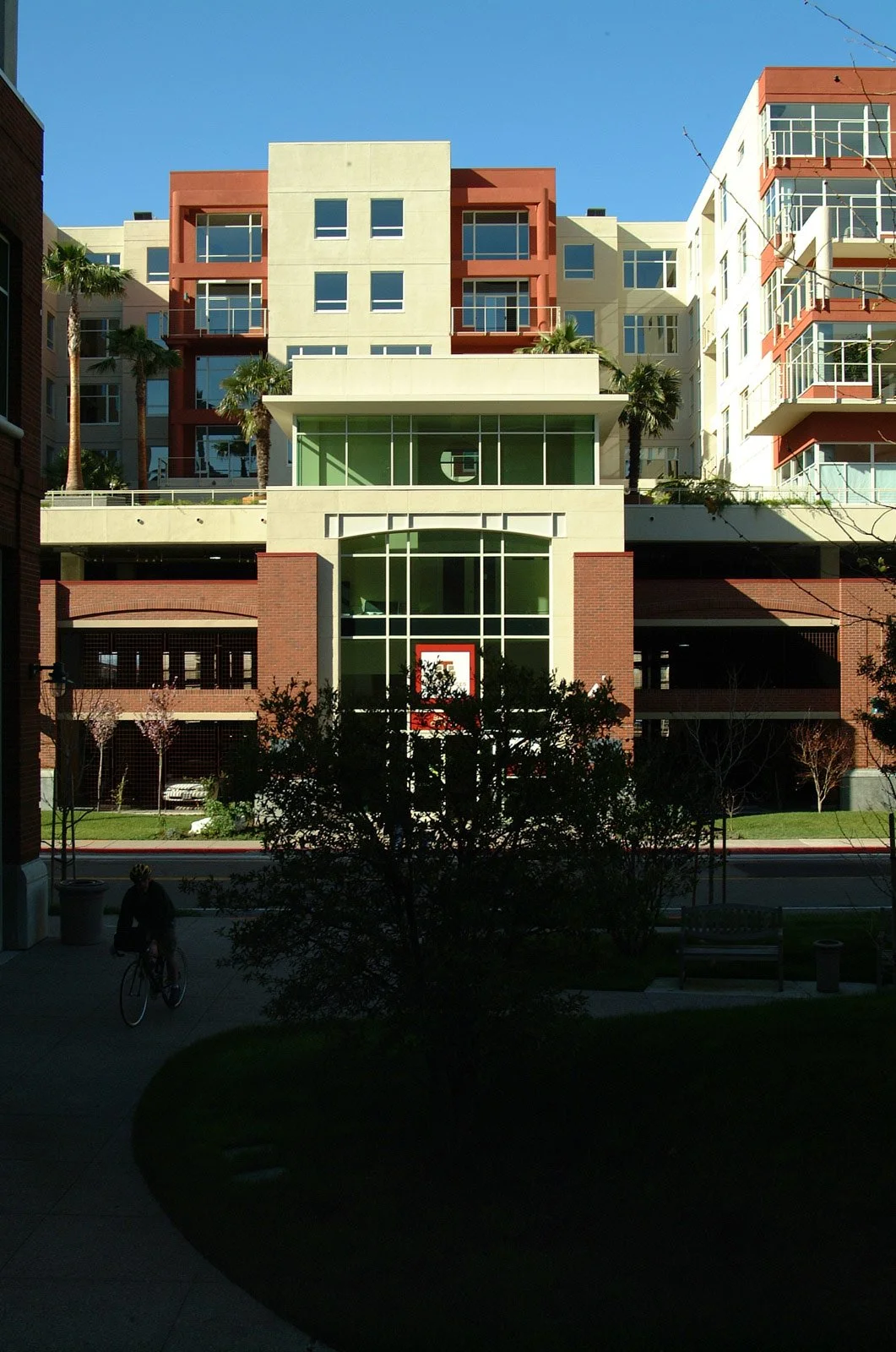 View of a modern apartment building with glass balconies, palm trees, and a basketball hoop in the foreground, under a clear blue sky.
