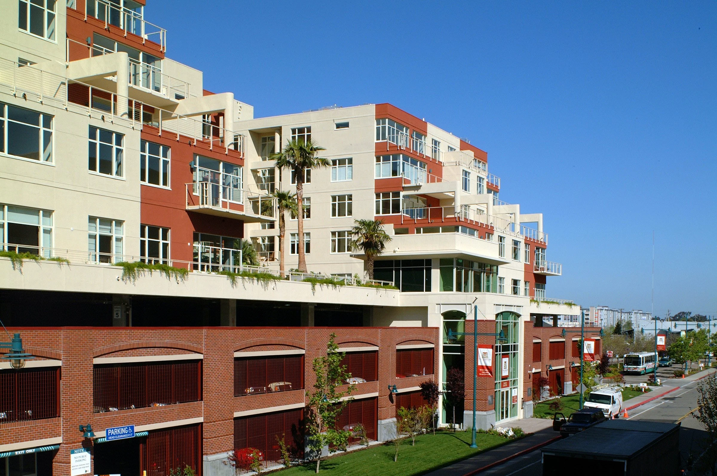 A modern multi-story apartment building with white and red exterior walls, large glass windows, and balconies. Palm trees are in front, and a street with parked cars and buses runs along the sidewalk.
