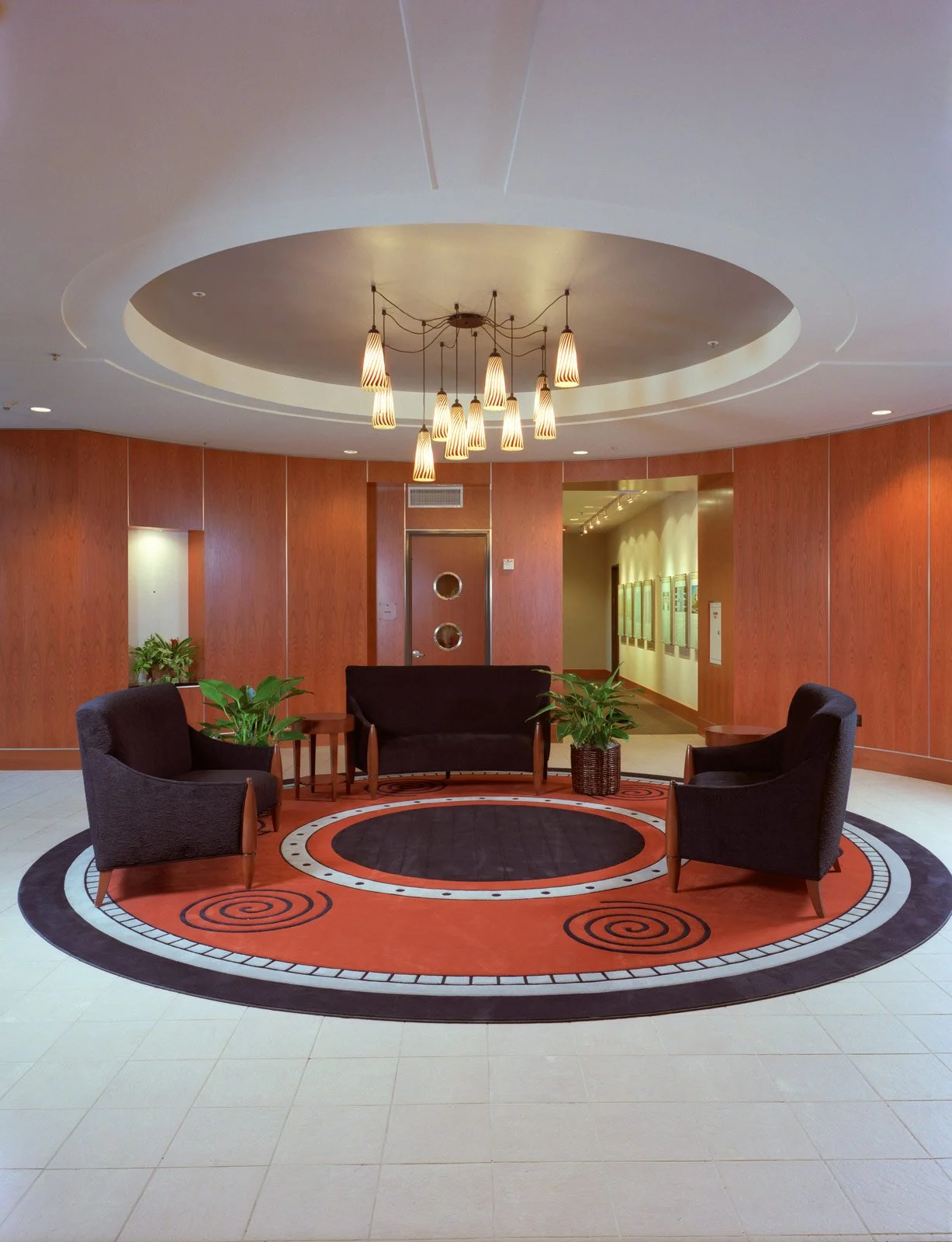 Modern hotel lobby with a circular patterned rug, three black upholstered chairs, two potted plants, and a large ceiling chandelier with hanging lights.