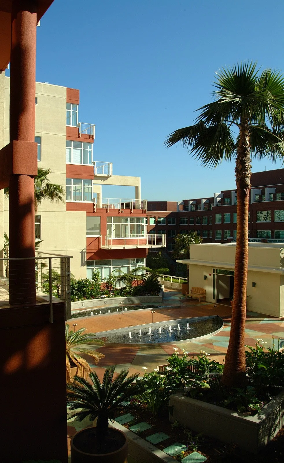 A view of a modern apartment complex courtyard with a fountain, palm trees, landscaping, and balconies in the background under a clear blue sky.