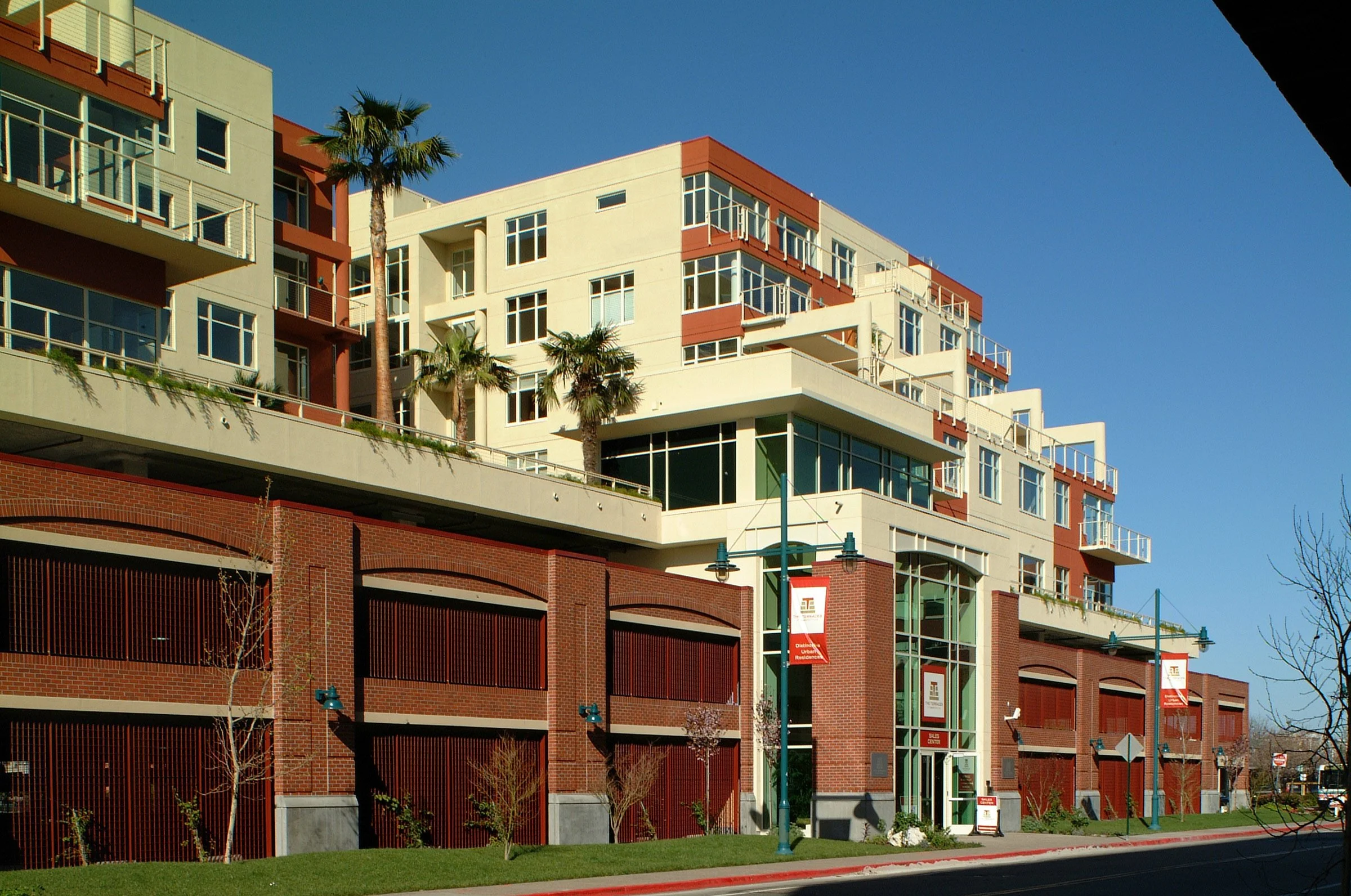 Modern multi-story apartment building with brick and beige exterior, large windows, palm trees, and a clear blue sky.