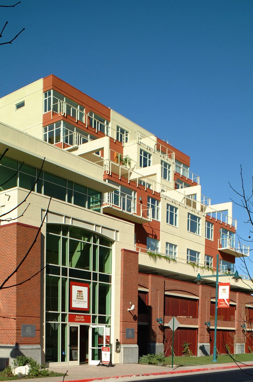 Modern multi-story residential building with brick and white exterior, large windows, balconies, and a storefront with a sign that reads 'The Terraces, Sales Center' at street level, under a clear blue sky.