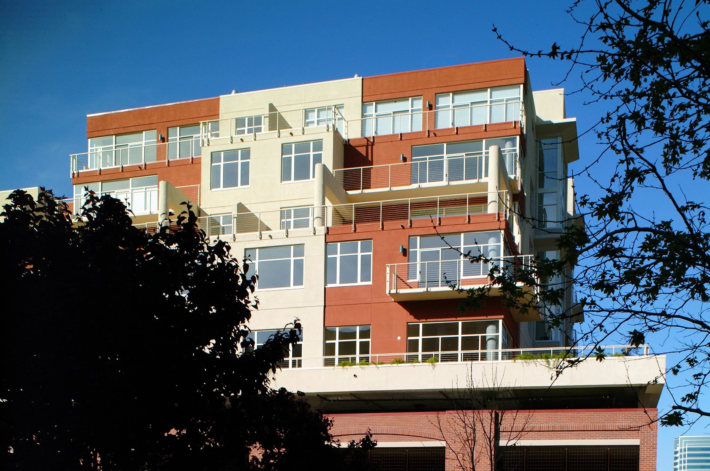 A modern multi-story apartment building with large windows and balconies, partly obscured by trees, against a clear blue sky.