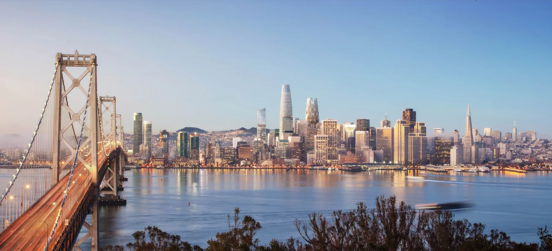 San Francisco skyline viewed across the bay with the Bay Bridge in the foreground and colorful city buildings, under a clear blue sky.