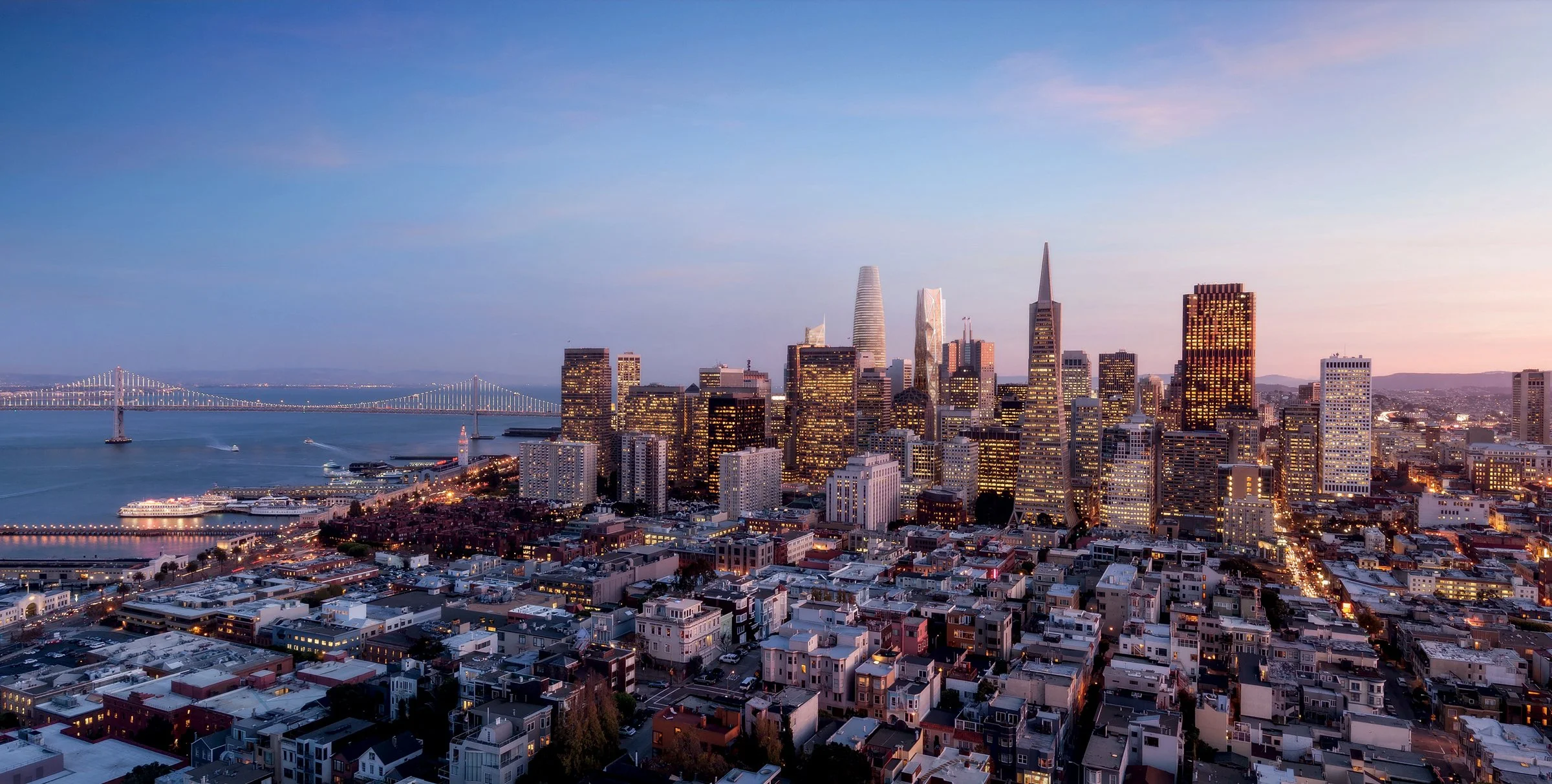San Francisco skyline at sunset with skyscrapers, Bay Bridge, and water with boats.