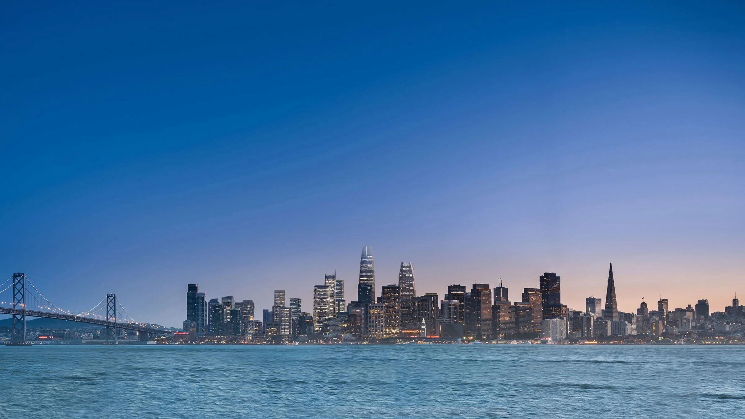 View of the San Francisco skyline at dusk with the Bay Bridge on the left, water in the foreground, and city buildings illuminated against a blue evening sky.