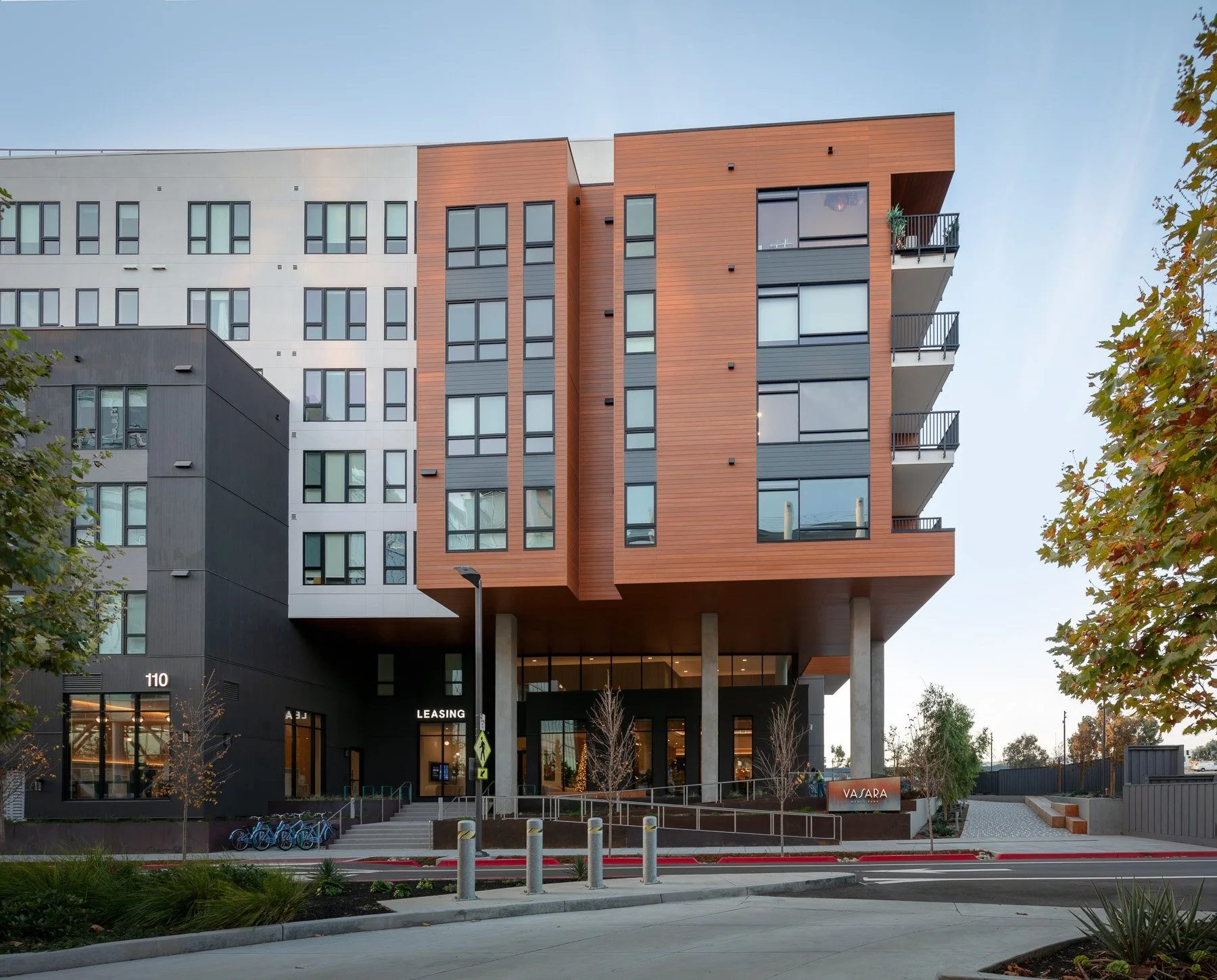 Modern multi-story apartment building with a black and brown facade, large windows, and balconies, located on a city street with trees and bicycle racks.